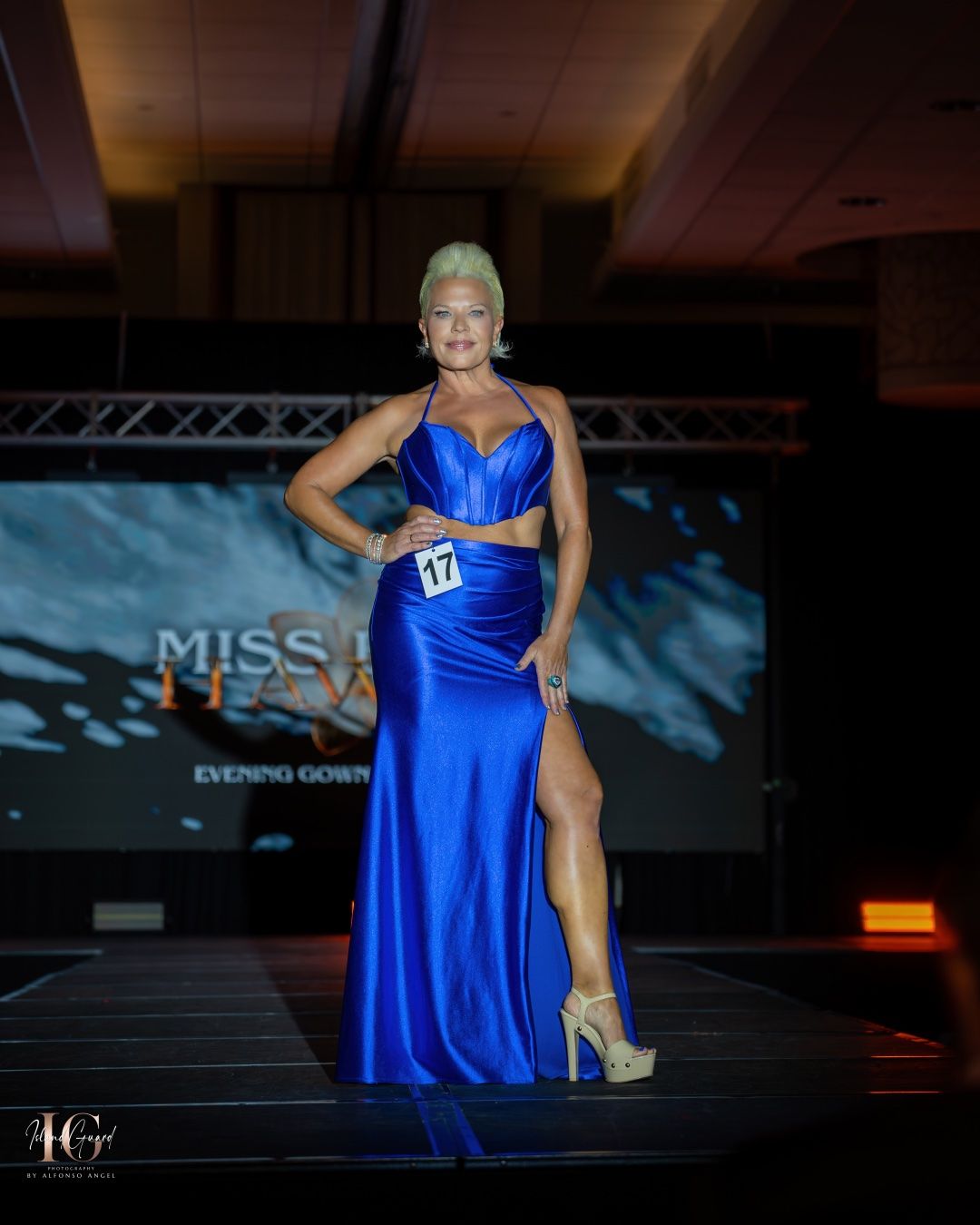 A pageant contestant in a vibrant royal blue evening gown with a high slit poses confidently on stage under dramatic lighting.