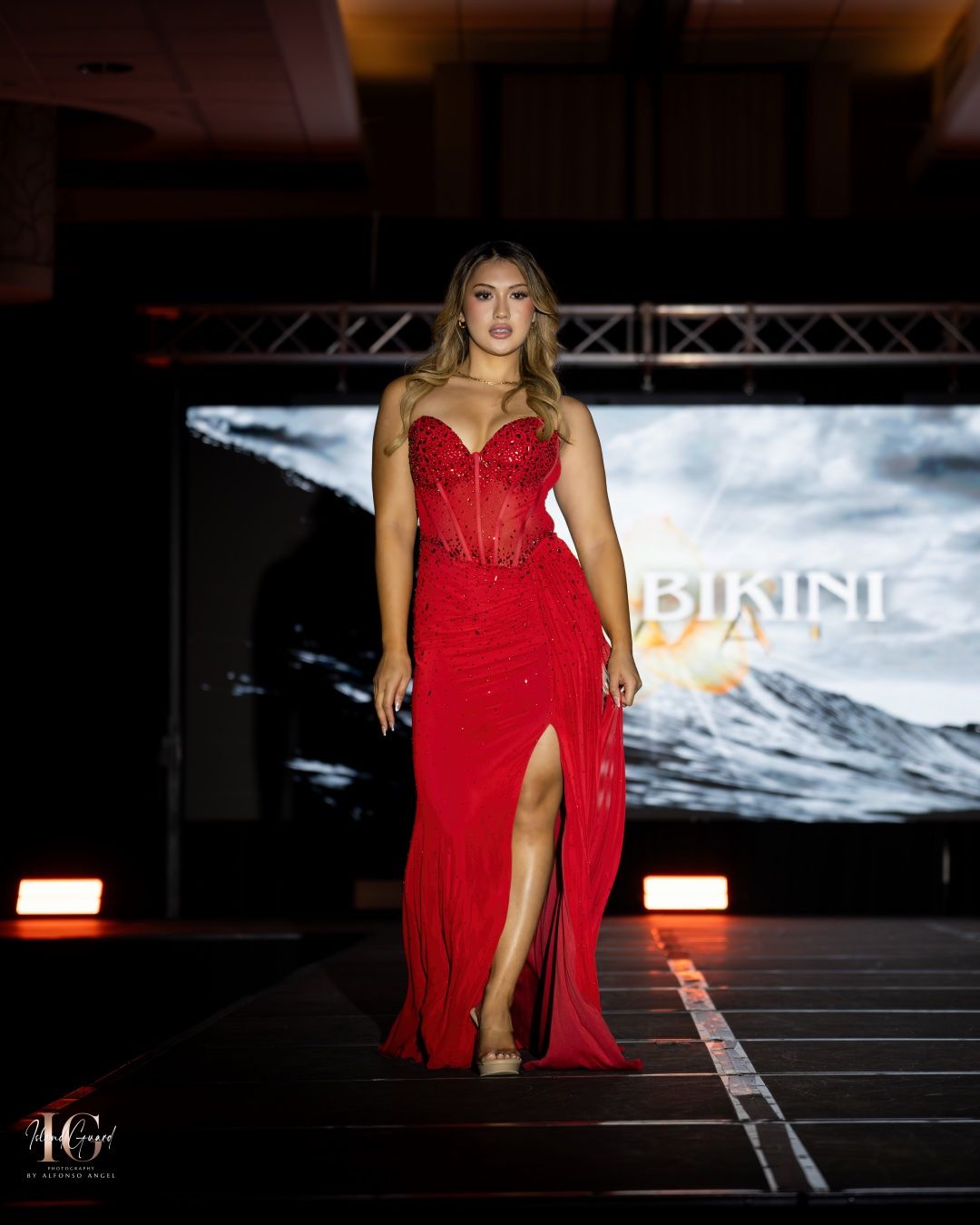 A model walks the runway in a stunning red evening gown with corset bodice and high slit at a fashion show.