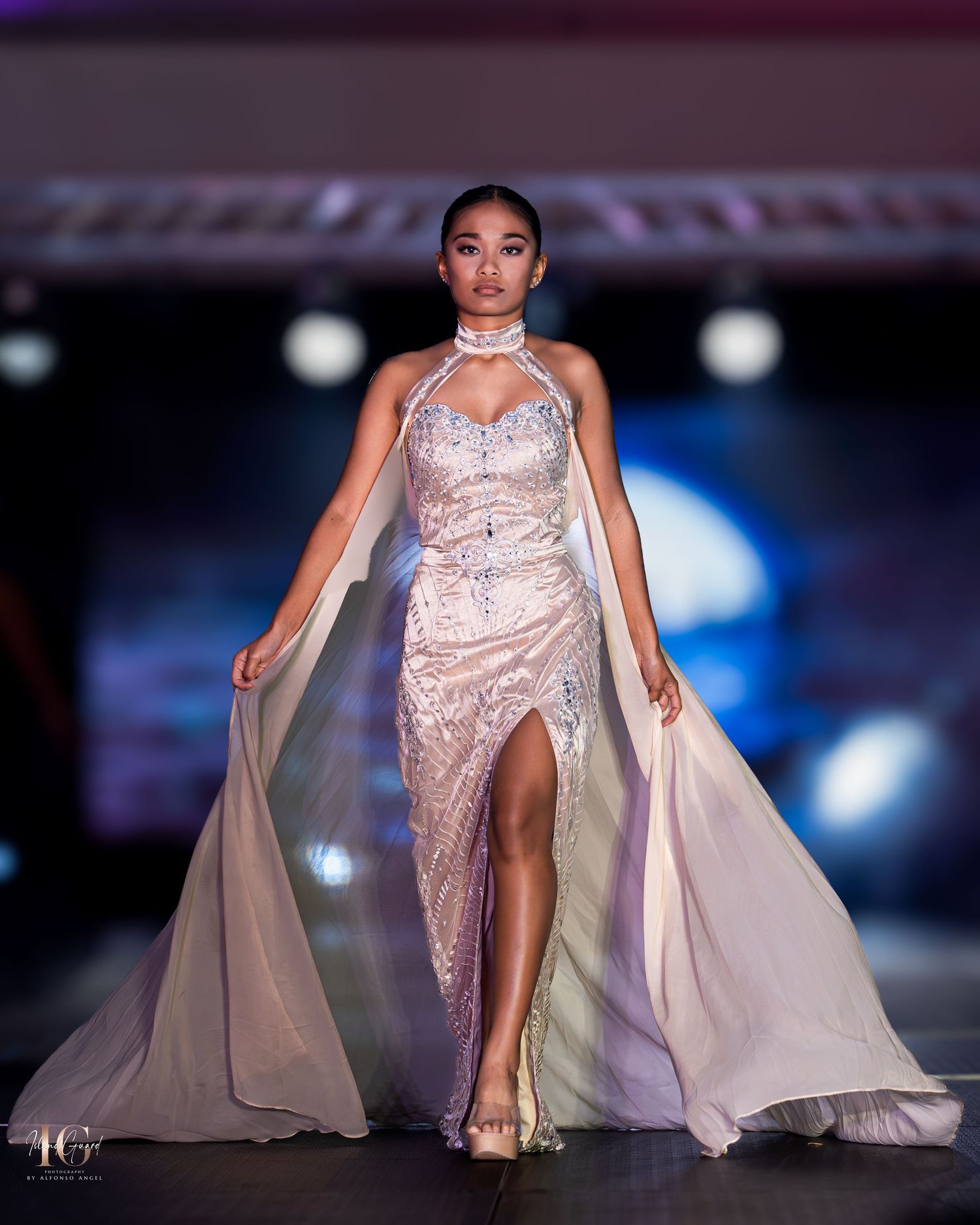 Model showcases elegant champagne-colored gown with cape and crystal embellishments on fashion runway.