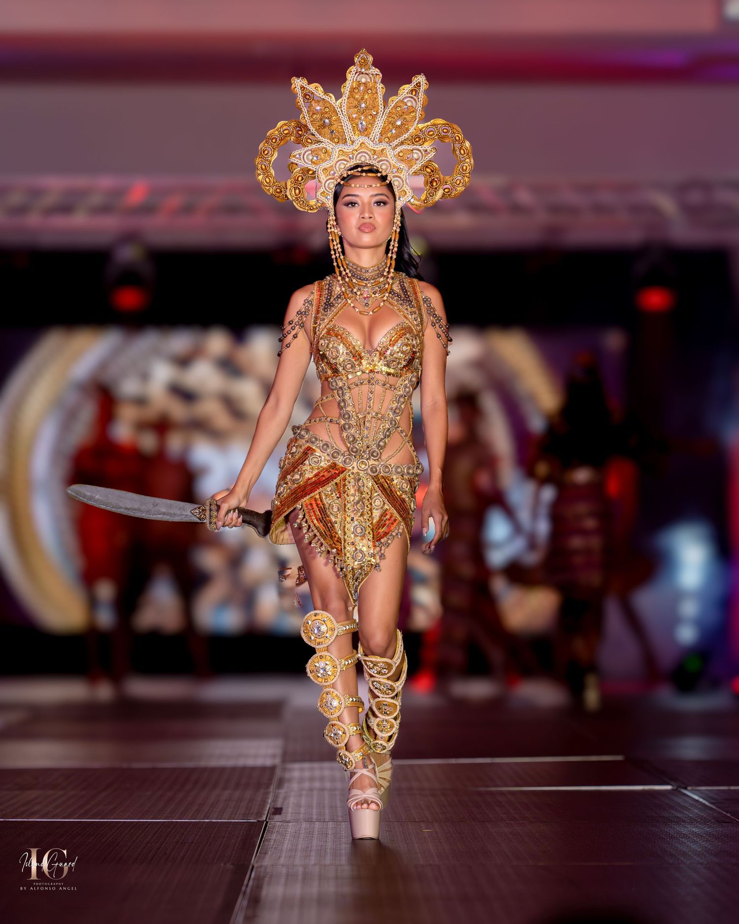 Fashion model wearing ornate gold warrior costume with sunburst crown headpiece on runway.
