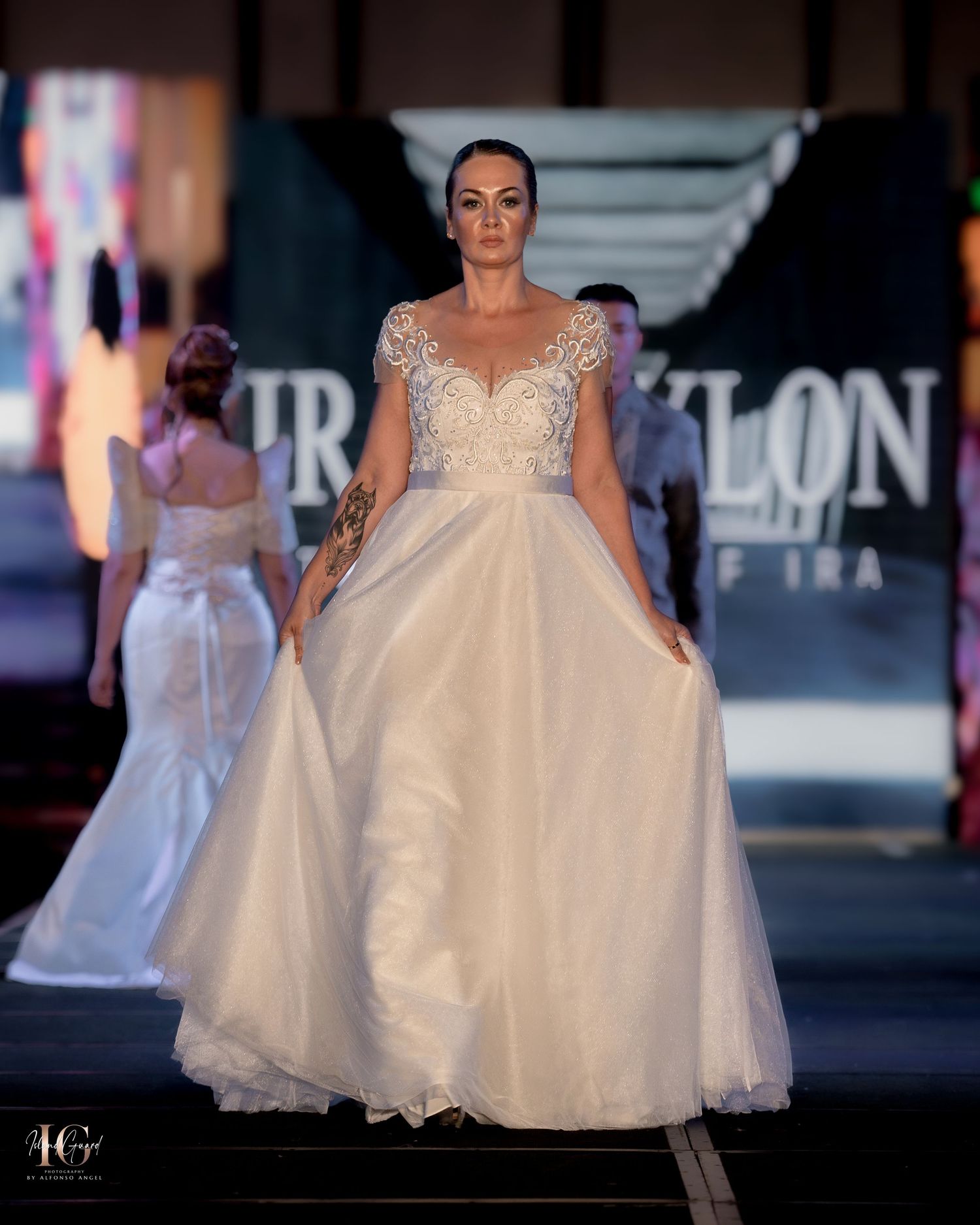 Models showcase collection of white formal gowns on illuminated runway with dark backdrop.