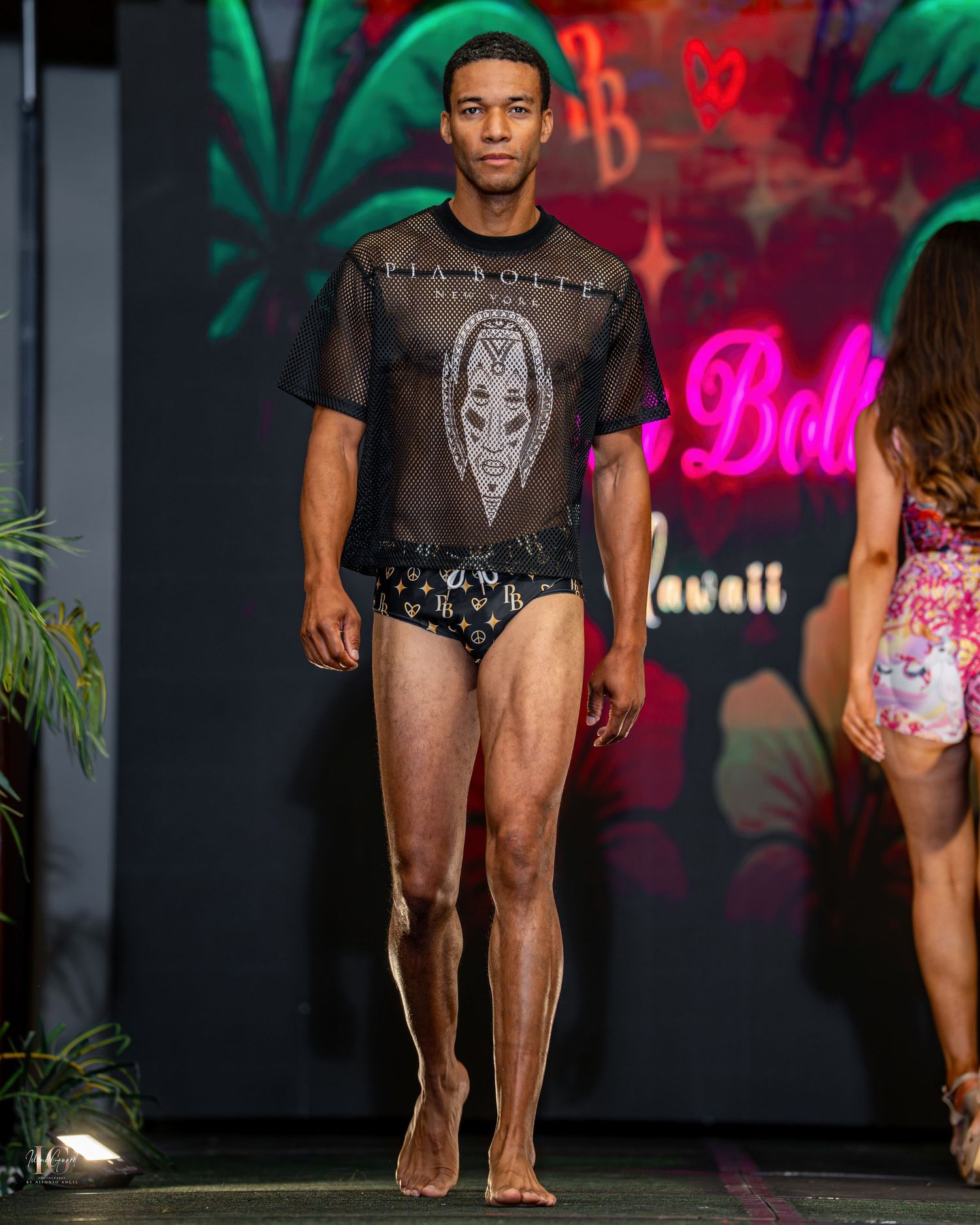 Model walks runway in sheer black graphic t-shirt and swim briefs at tropical-themed fashion show with neon lighting.
