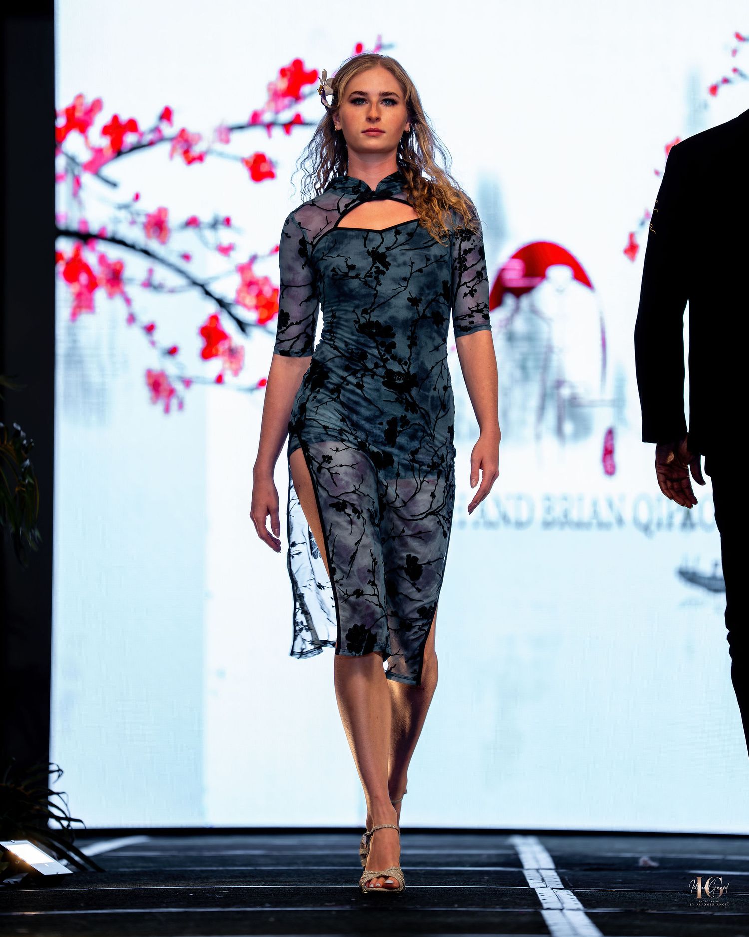A model walks the runway in a dark patterned dress during a fashion show with cherry blossom decor in the background.