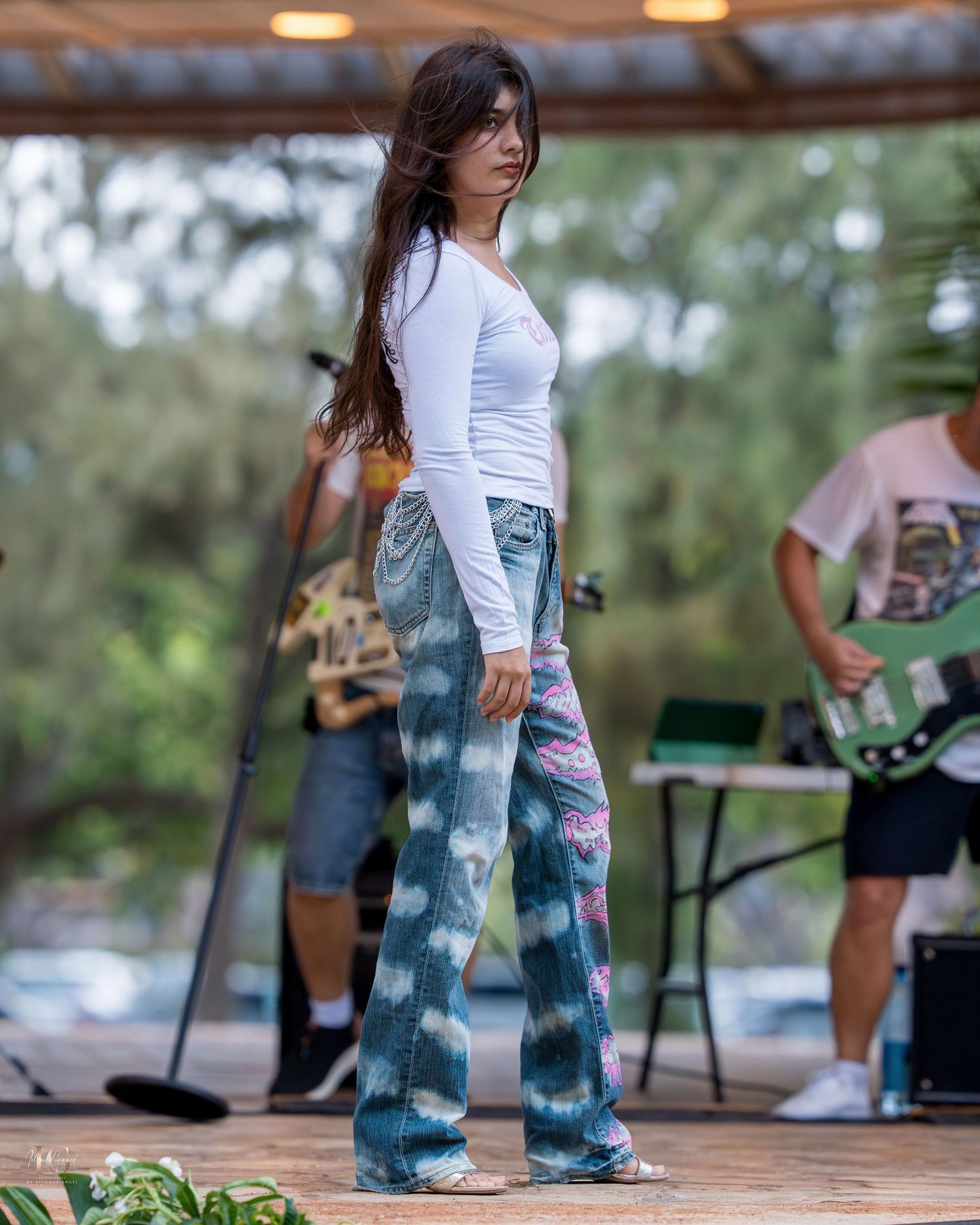 A person wearing tie-dyed jeans and white top stands outdoors with blurred background of trees and other people.