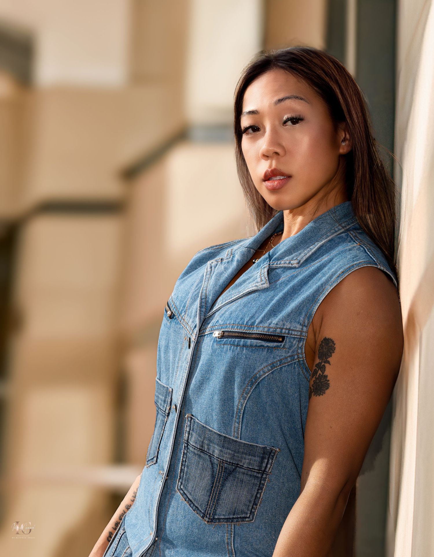 Person in denim jacket vest poses against a neutral colored wall in natural lighting.