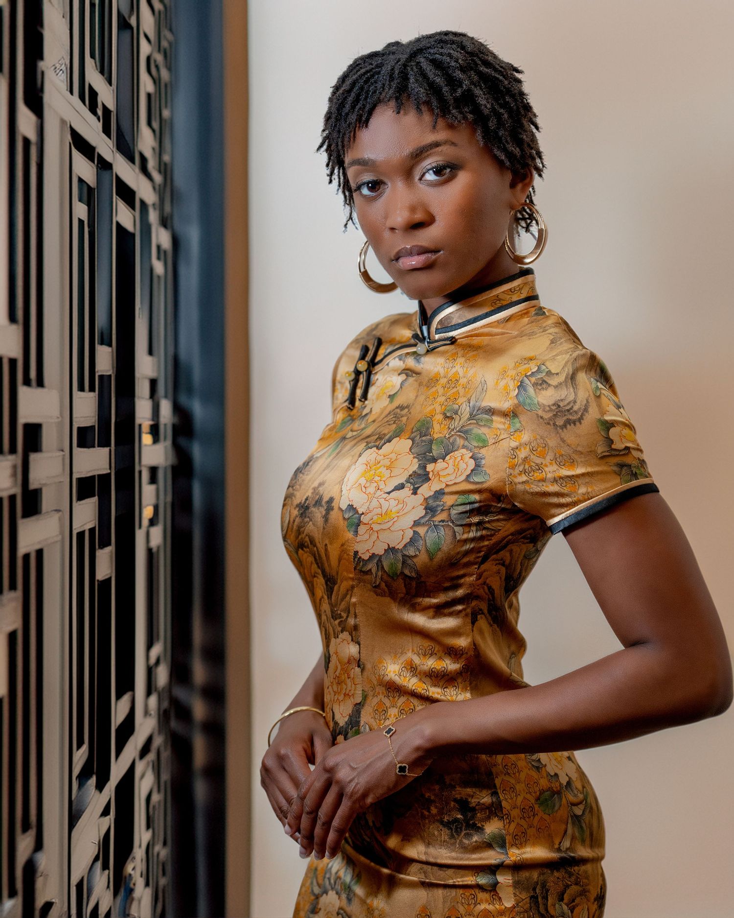 A person wearing a traditional gold Chinese cheongsam dress with floral pattern poses against a modern patterned wall.