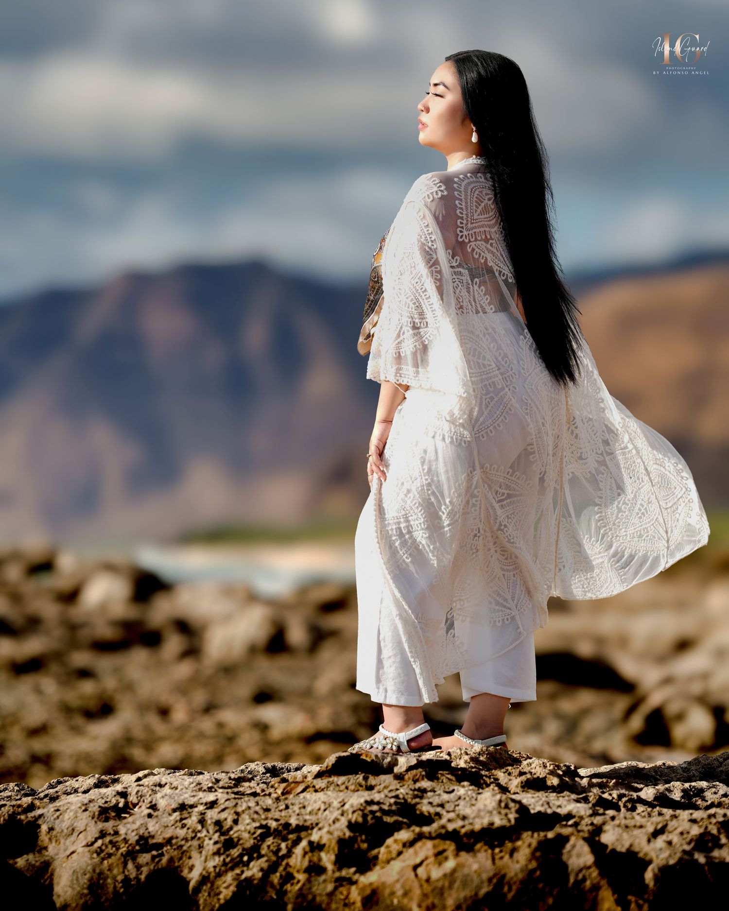 A person in a flowing white lace dress stands gracefully on rocky terrain against a cloudy mountain landscape.