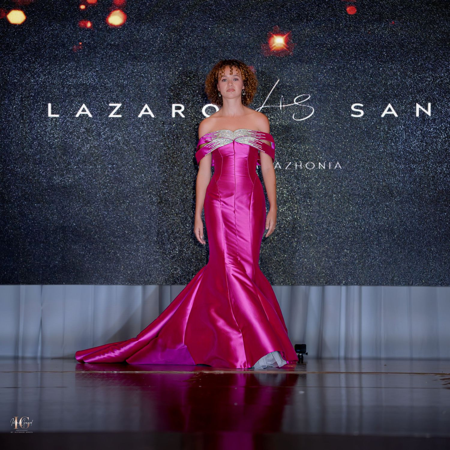 Sleek hot pink mermaid gown with off-shoulder detail shown on illuminated runway.
