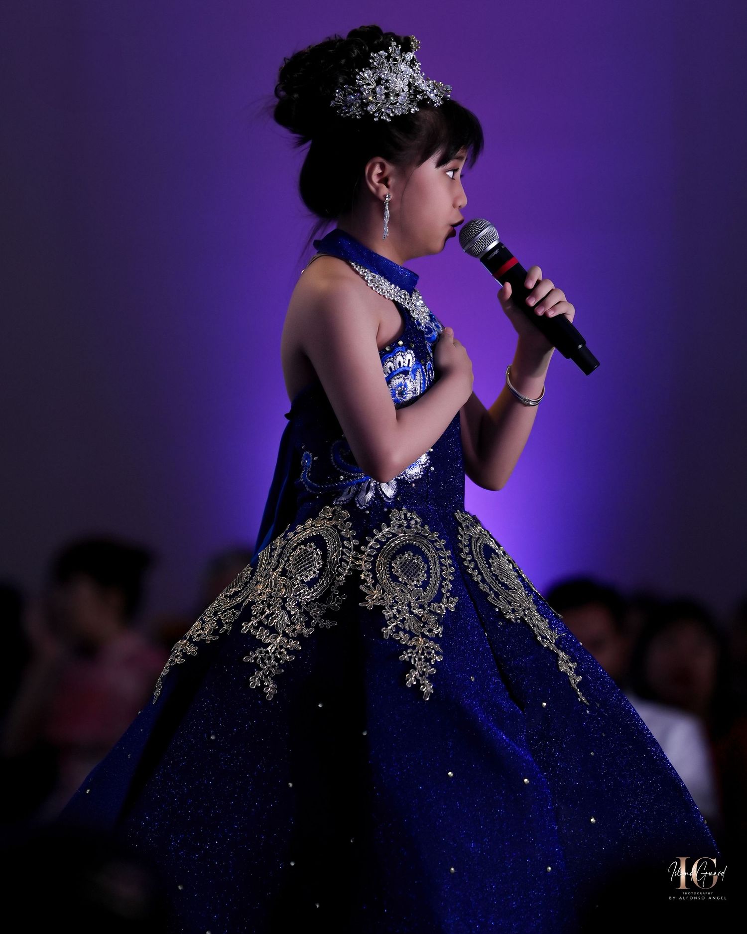 Young performer in navy blue ballgown with gold embellishments sings on stage.