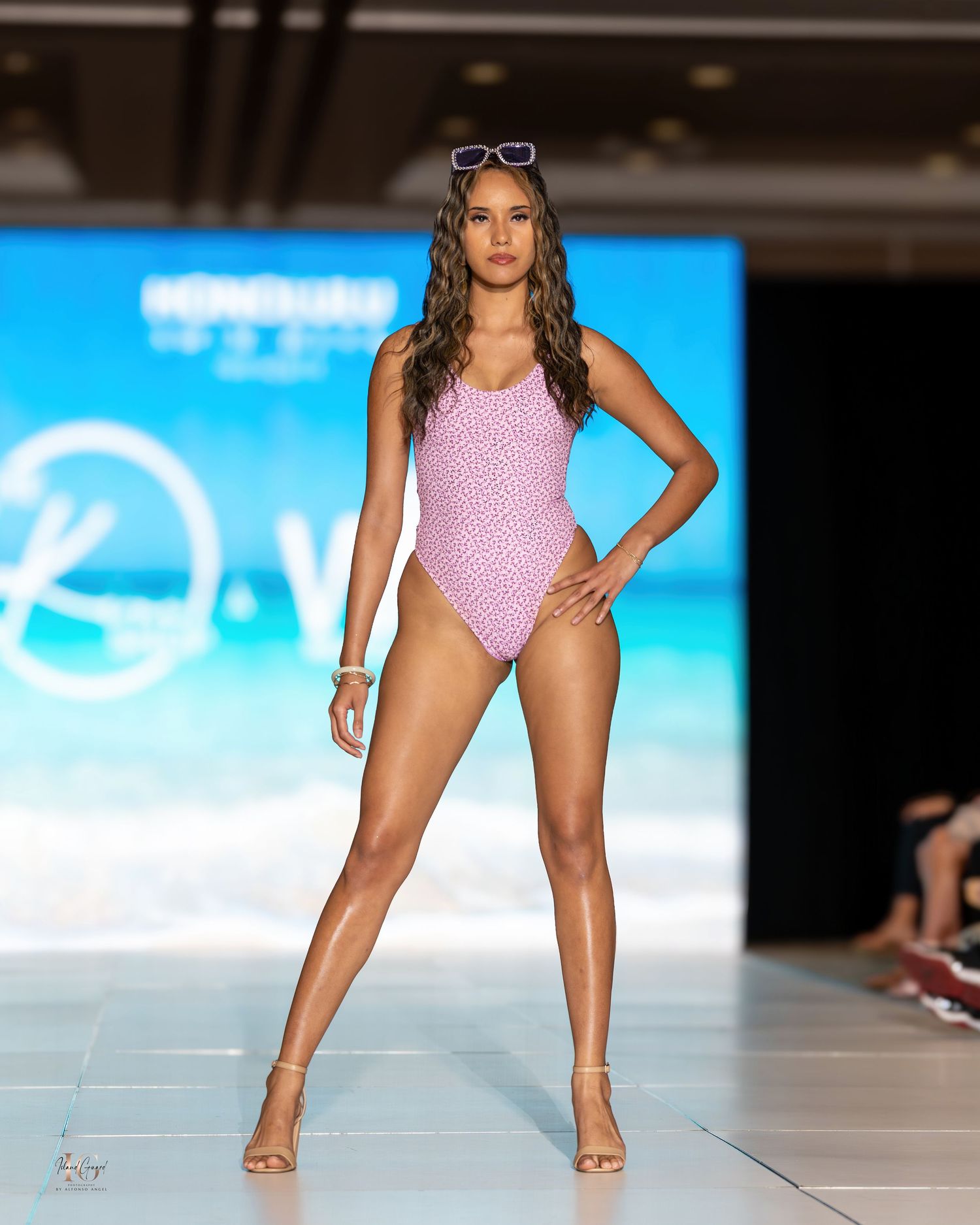 Model strides down runway in pink swimsuit during fashion show.