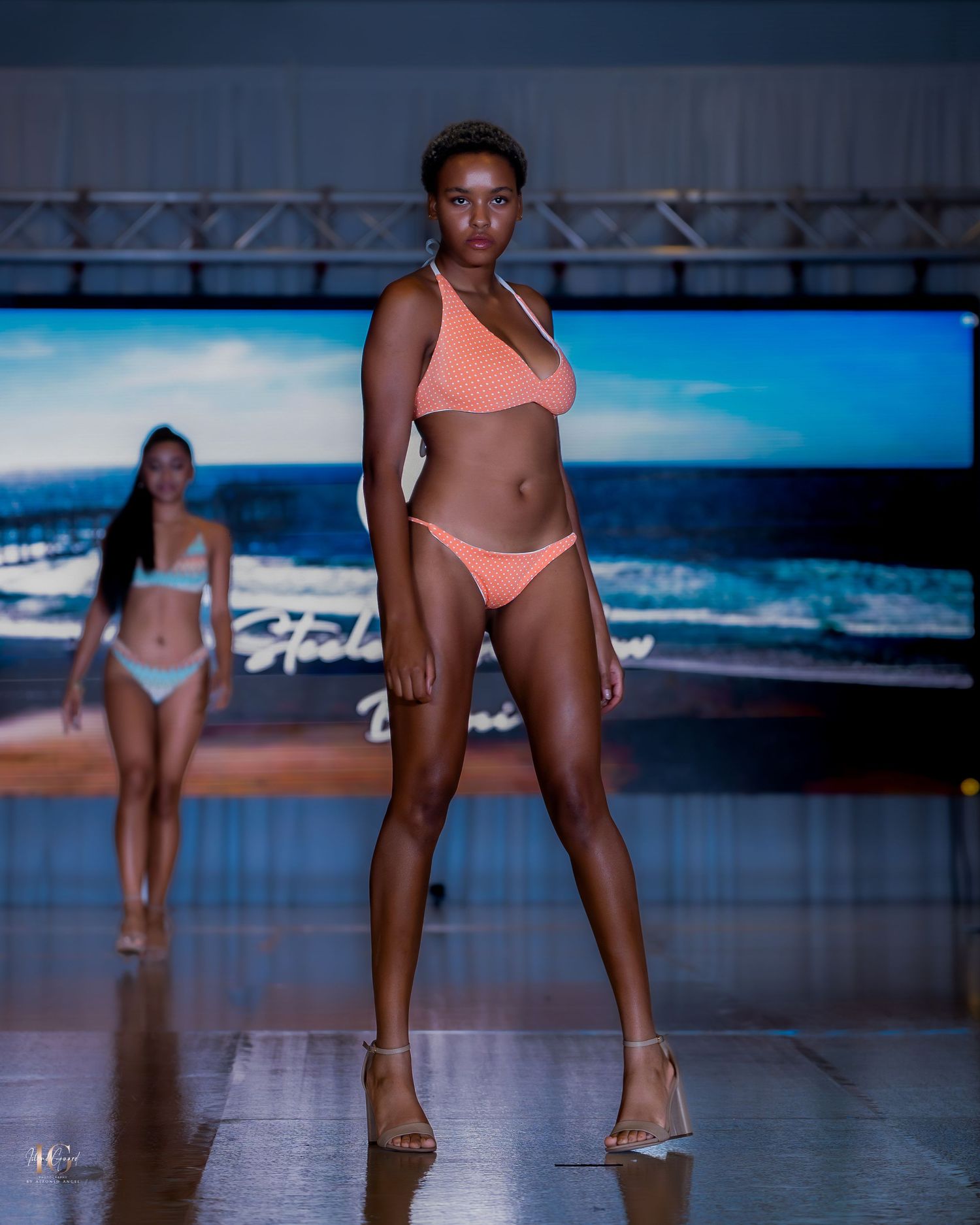Models walk the runway in swimwear during a fashion show against a blue-lit backdrop.