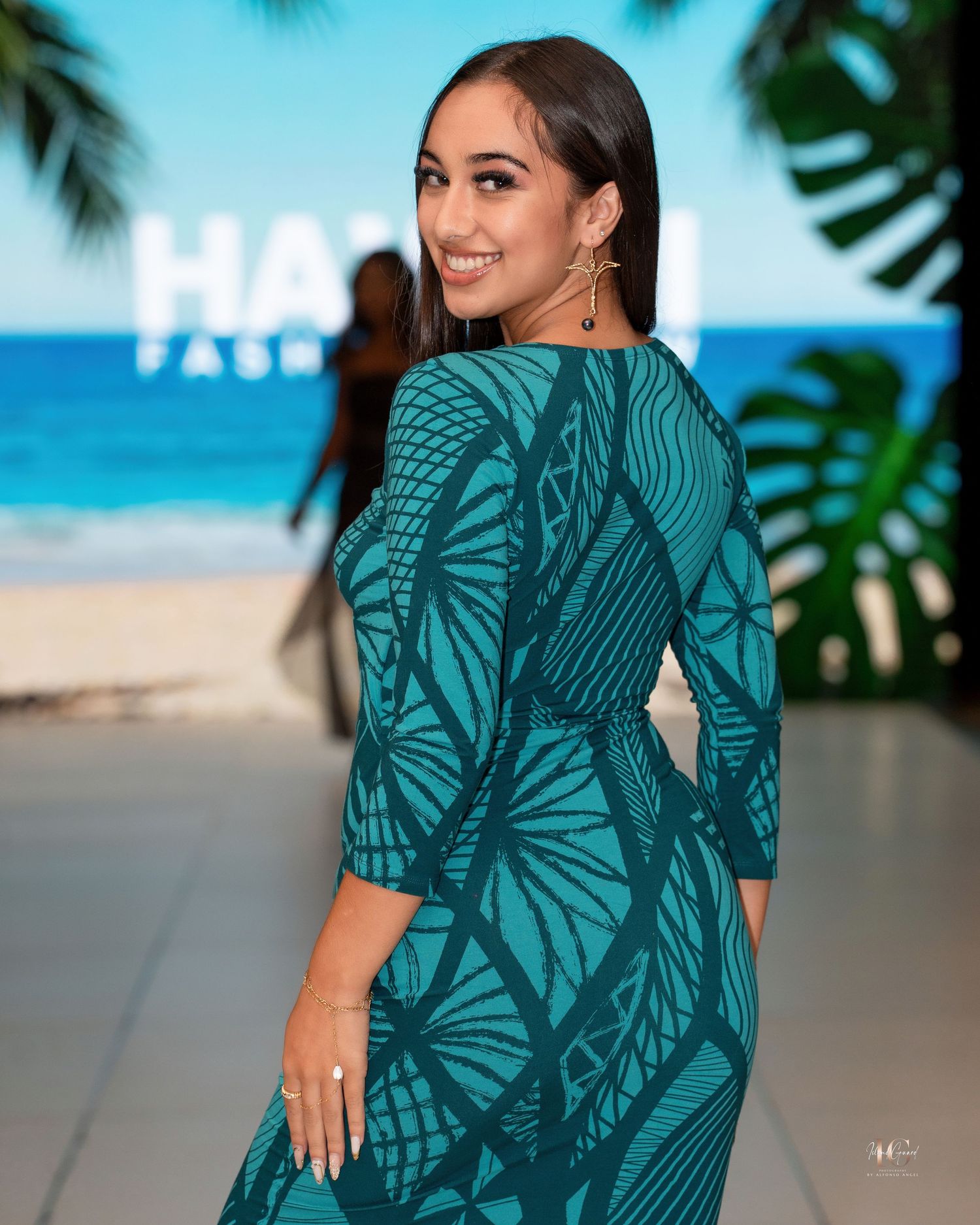 Woman in a tropical print teal dress poses against an ocean backdrop with palm fronds and beach scenery.