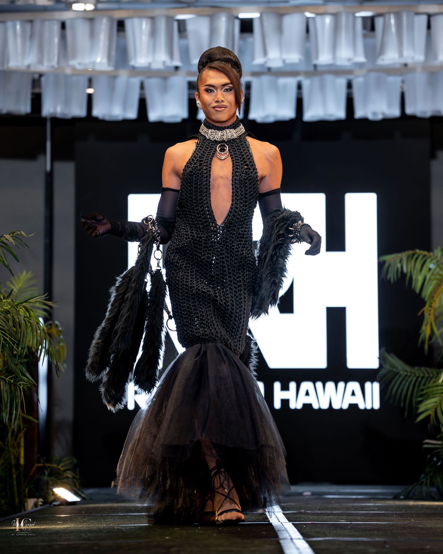 A model walks the runway in a dramatic black tulle gown with structured bodice at a fashion show in Hawaii.
