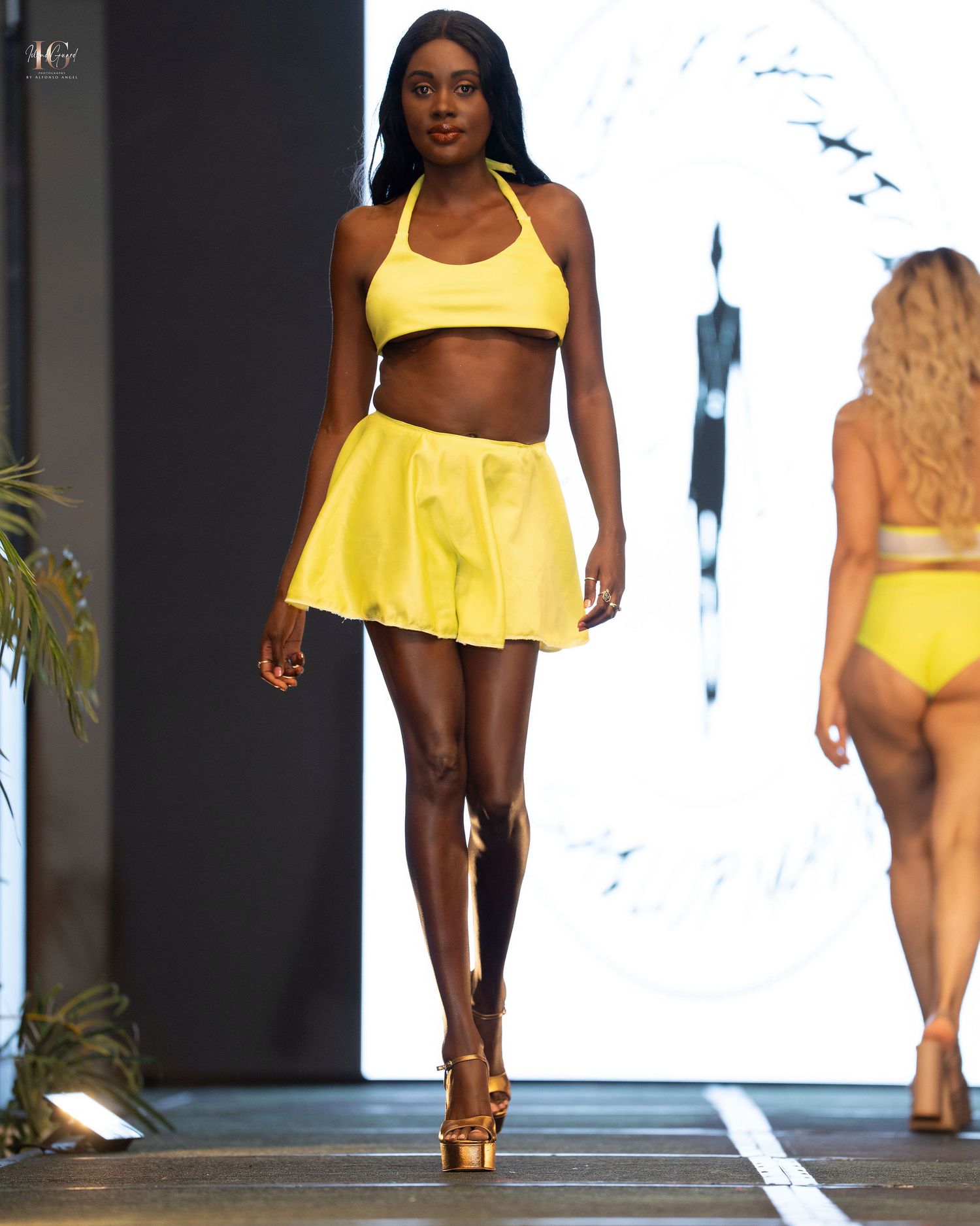 Models walk the runway in bright yellow swimwear and resort wear during a summer fashion show presentation.