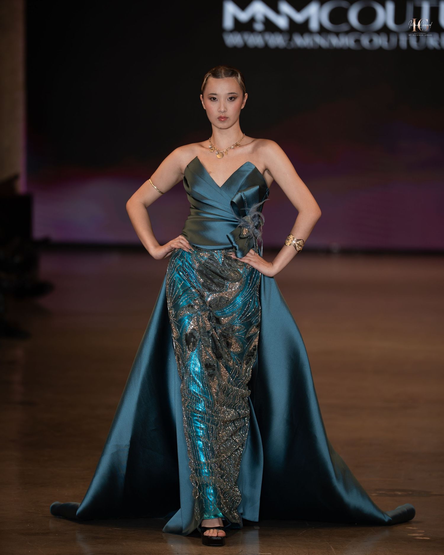 Model walks runway in teal evening gown with dramatic cape and metallic embellishments at fashion show.
