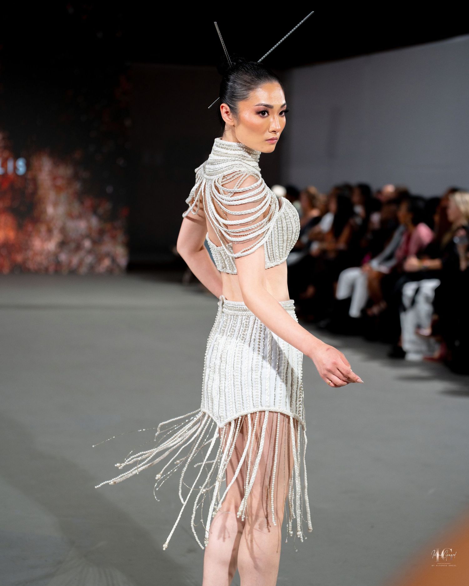 A model walks the runway wearing a white fringed dress with sheer panels and strappy details at a fashion show.