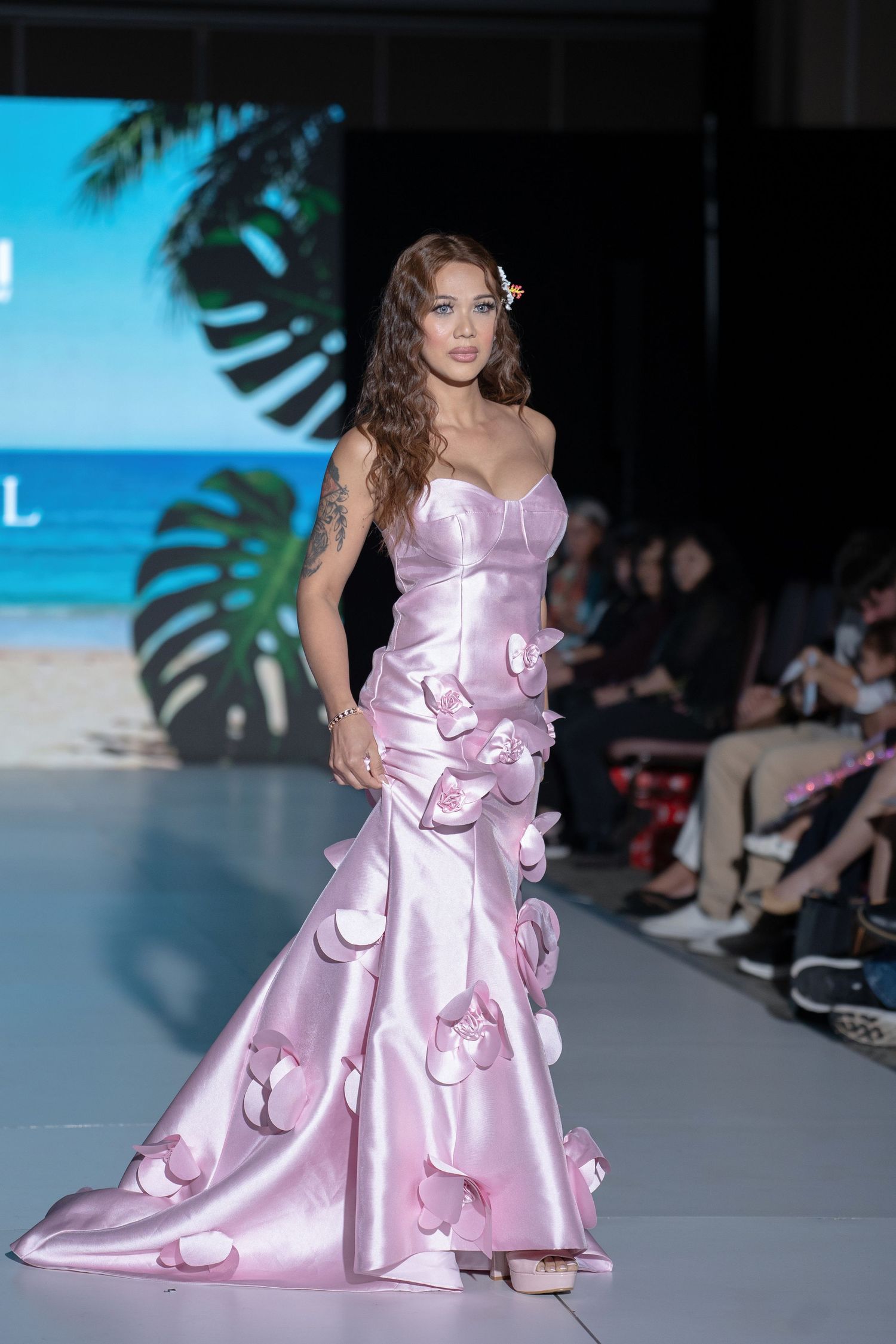 Model walks runway in flowing pink satin gown with butterfly appliques at luxury fashion show.