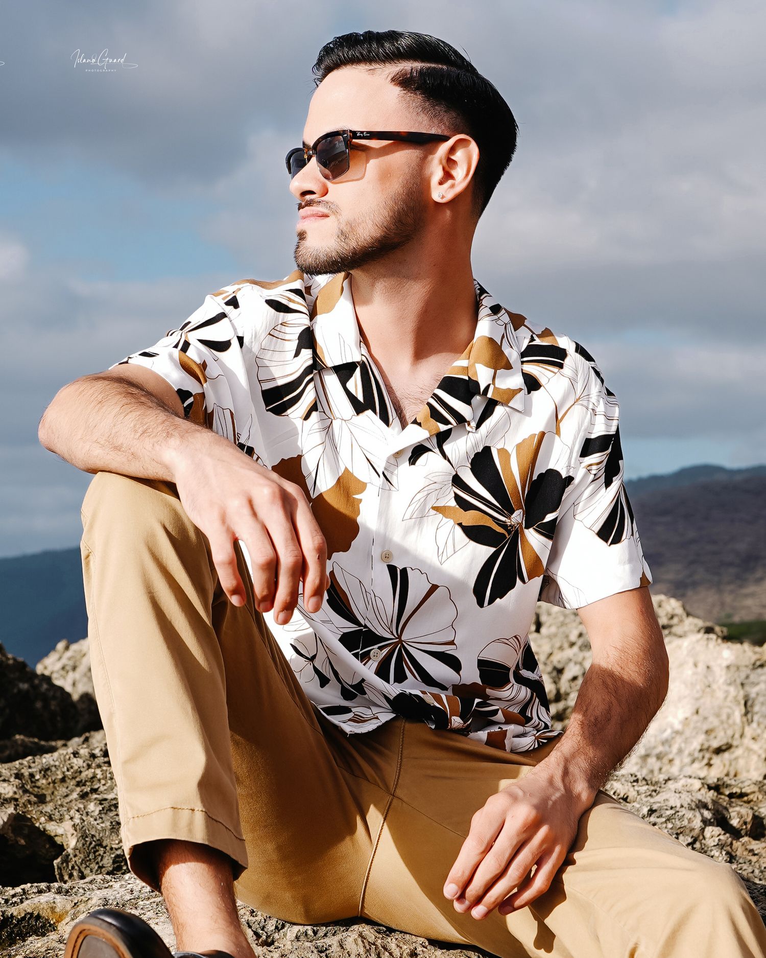 Person in floral print shirt and khaki shorts relaxing outdoors against mountainous desert landscape on sunny day.