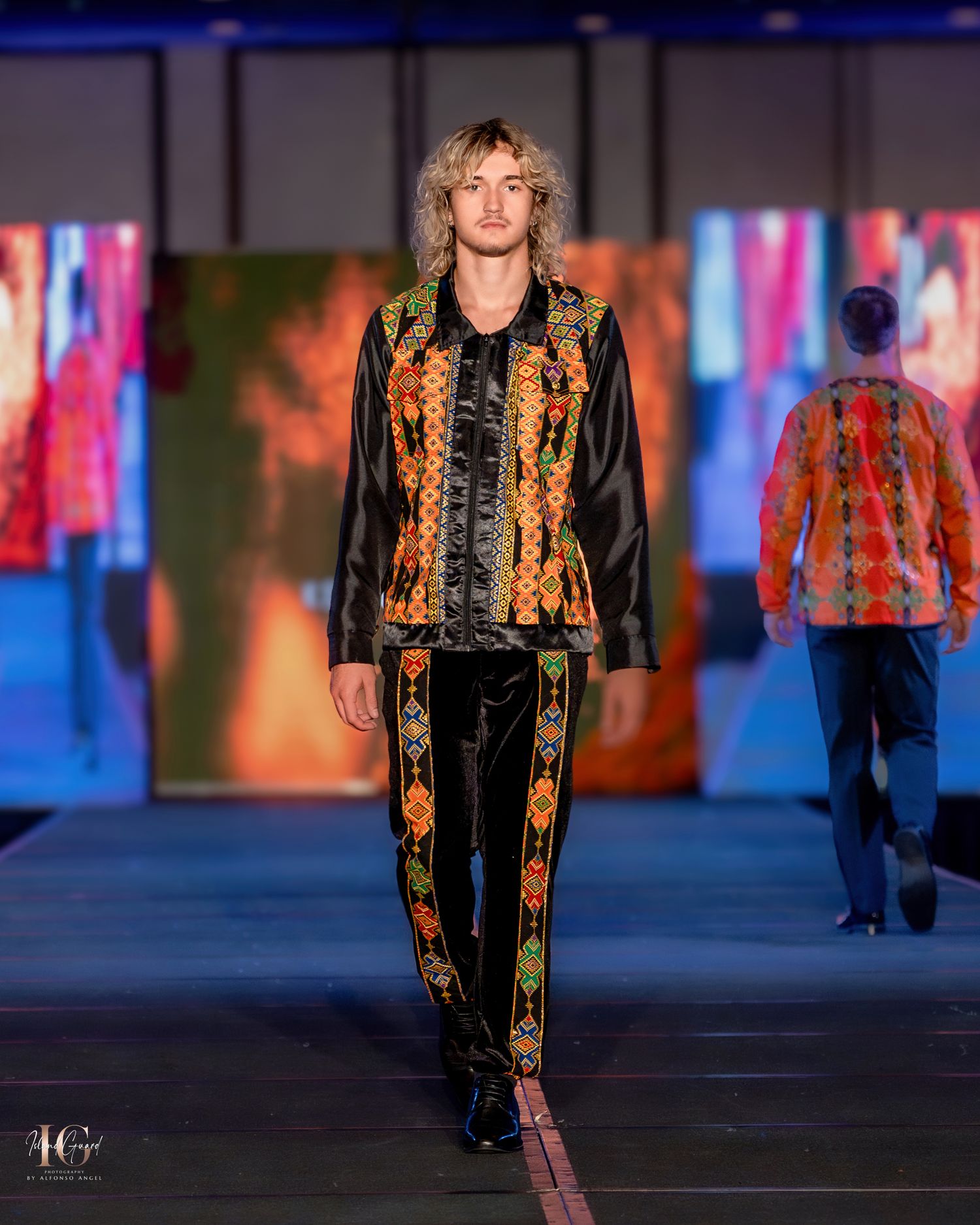 Fashion model walks runway wearing black and gold patterned jacket during evening fashion show.