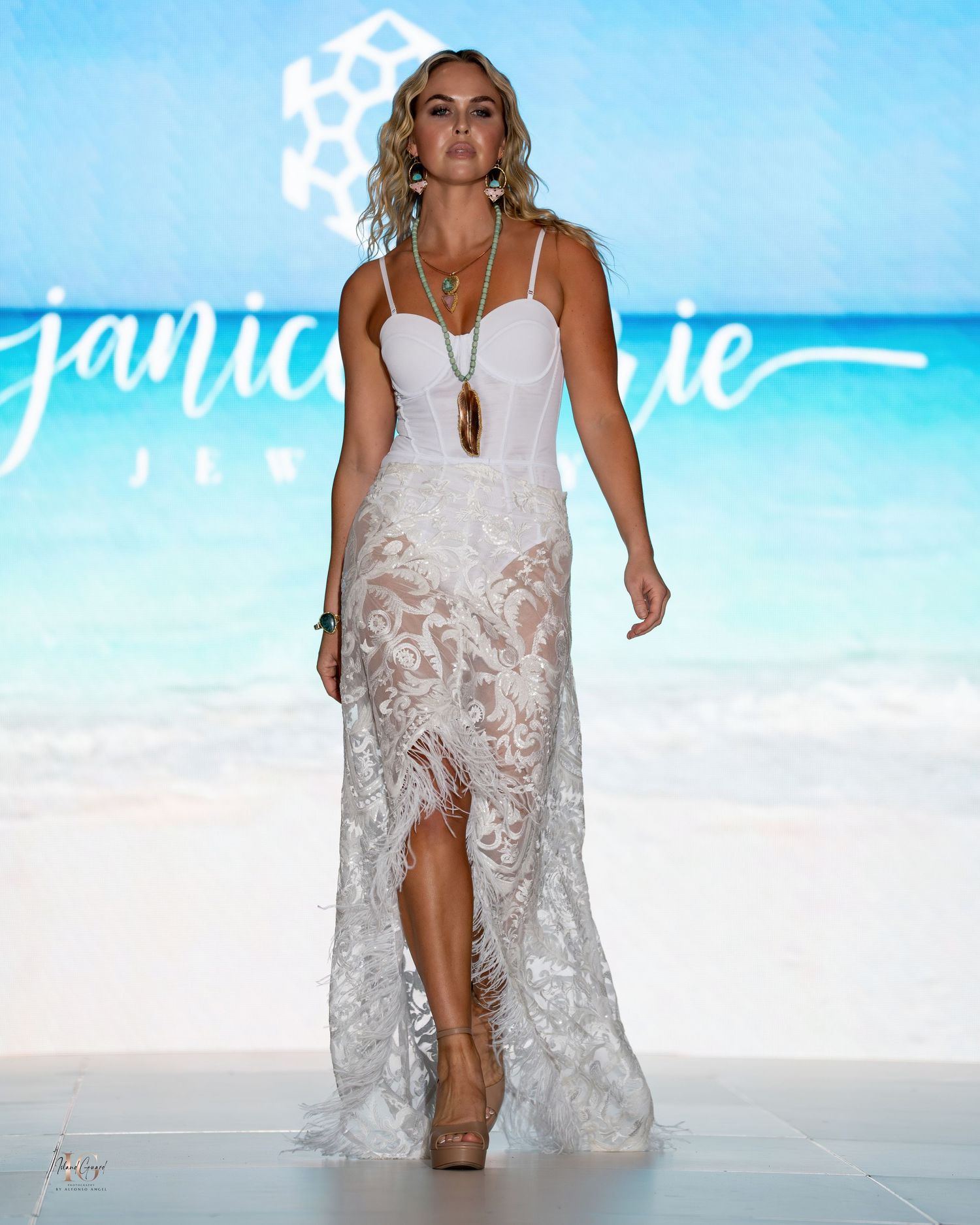 Model in white lace dress with high slit walks runway at beach-themed fashion show.