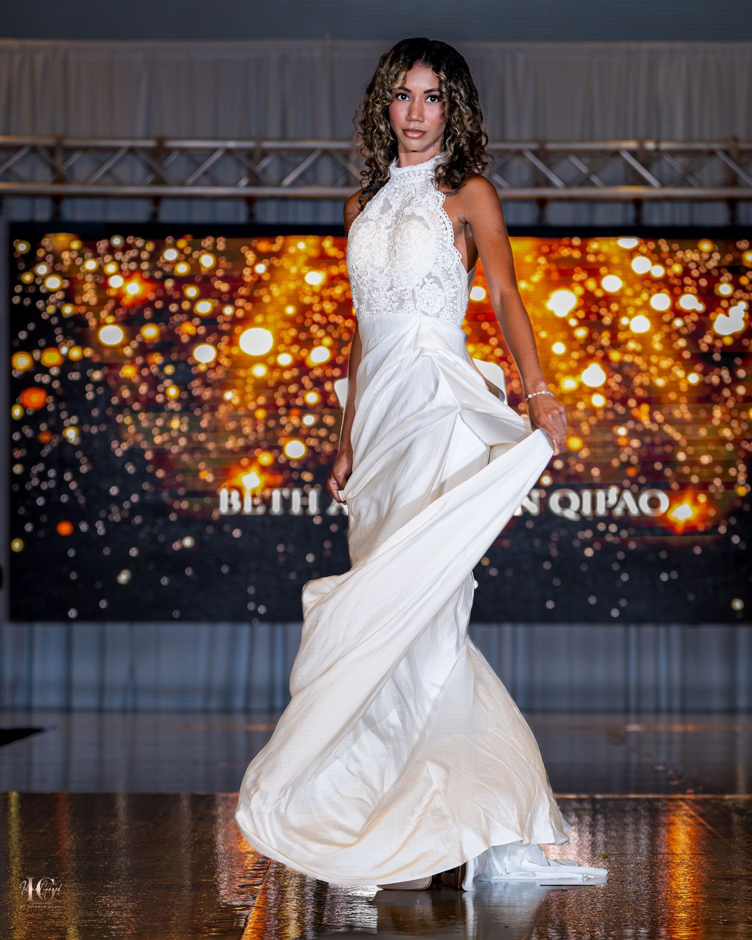 Model twirls in flowing white halter gown against golden bokeh backdrop on fashion runway.