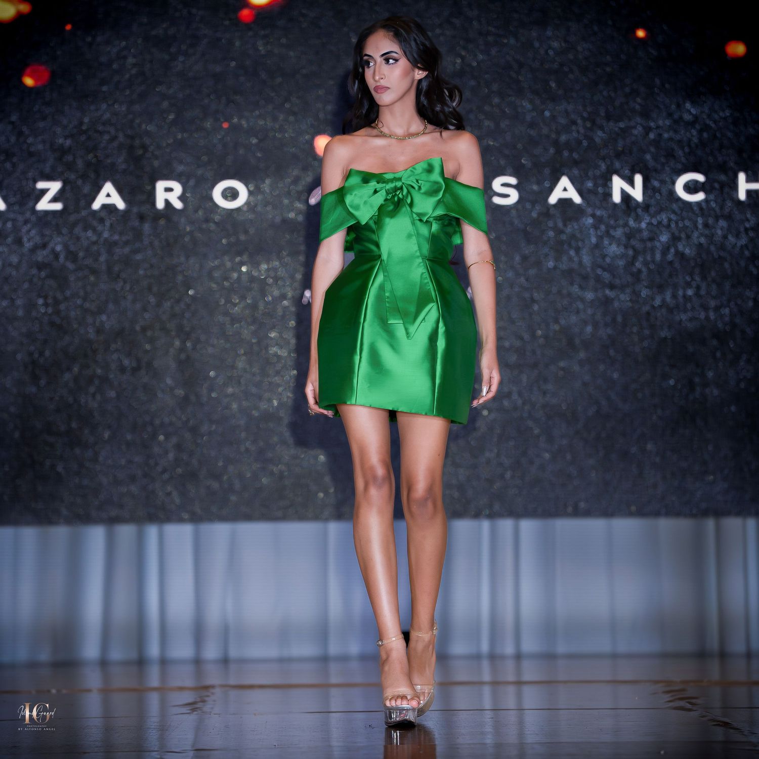 Model presents emerald green off-shoulder mini dress on dark runway.