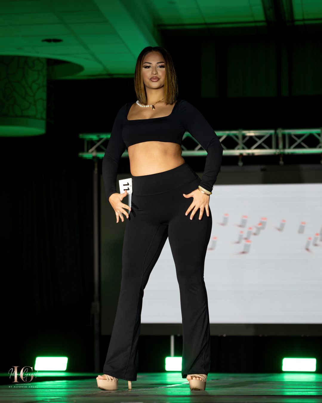 Model poses in black activewear set with crop top and fitted pants on fashion runway with green lighting.