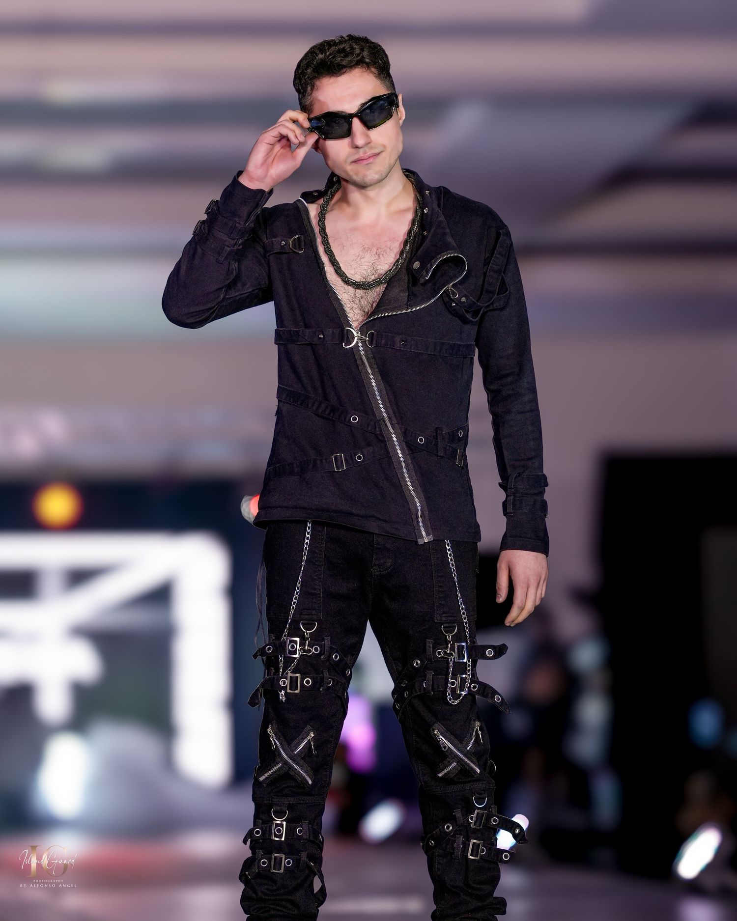 Model in edgy black outfit with sunglasses and decorative details on fashion runway.