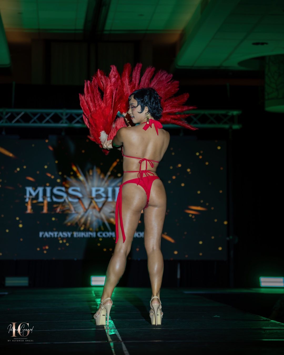 A performer in a red costume with dramatic red feather headpiece dances on stage at a fantasy show event.
