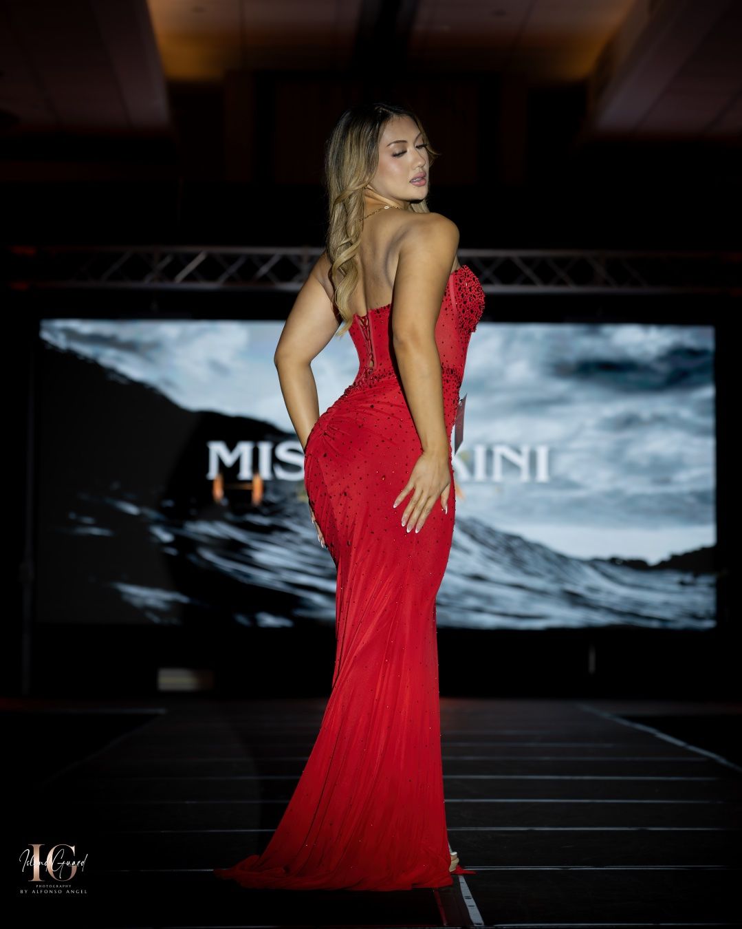 A model walks the runway in a form-fitting red evening gown during a fashion show with dramatic lighting and backdrop.