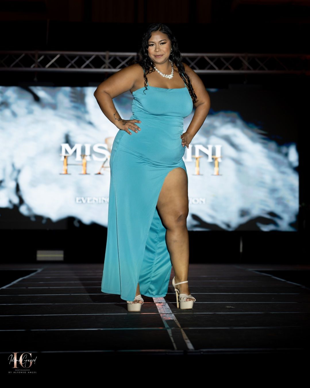 A model in a turquoise evening gown with thigh slit poses confidently on a fashion runway with stage lighting.
