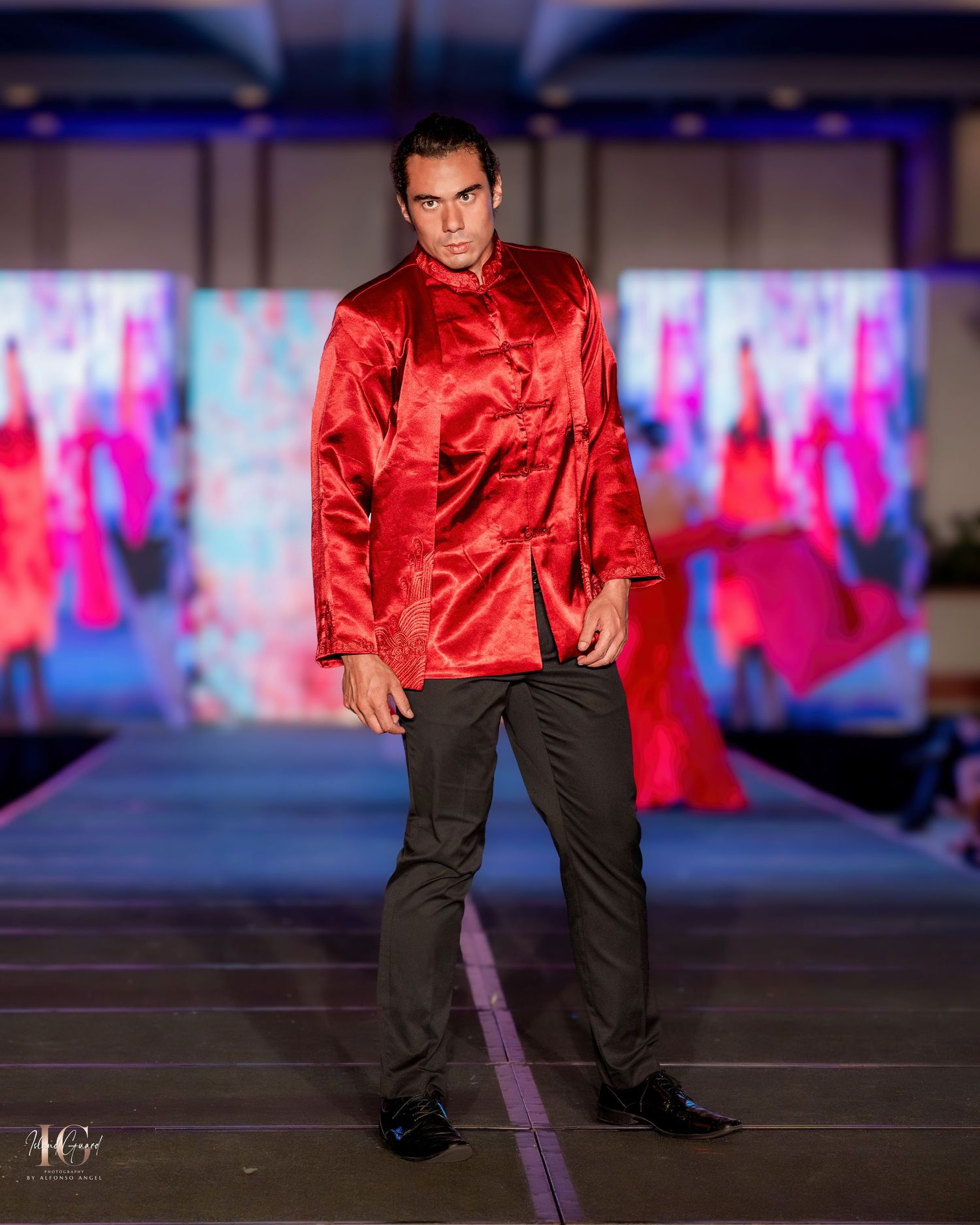 Model wearing red silk top and black pants performs on fashion runway with colored lighting.