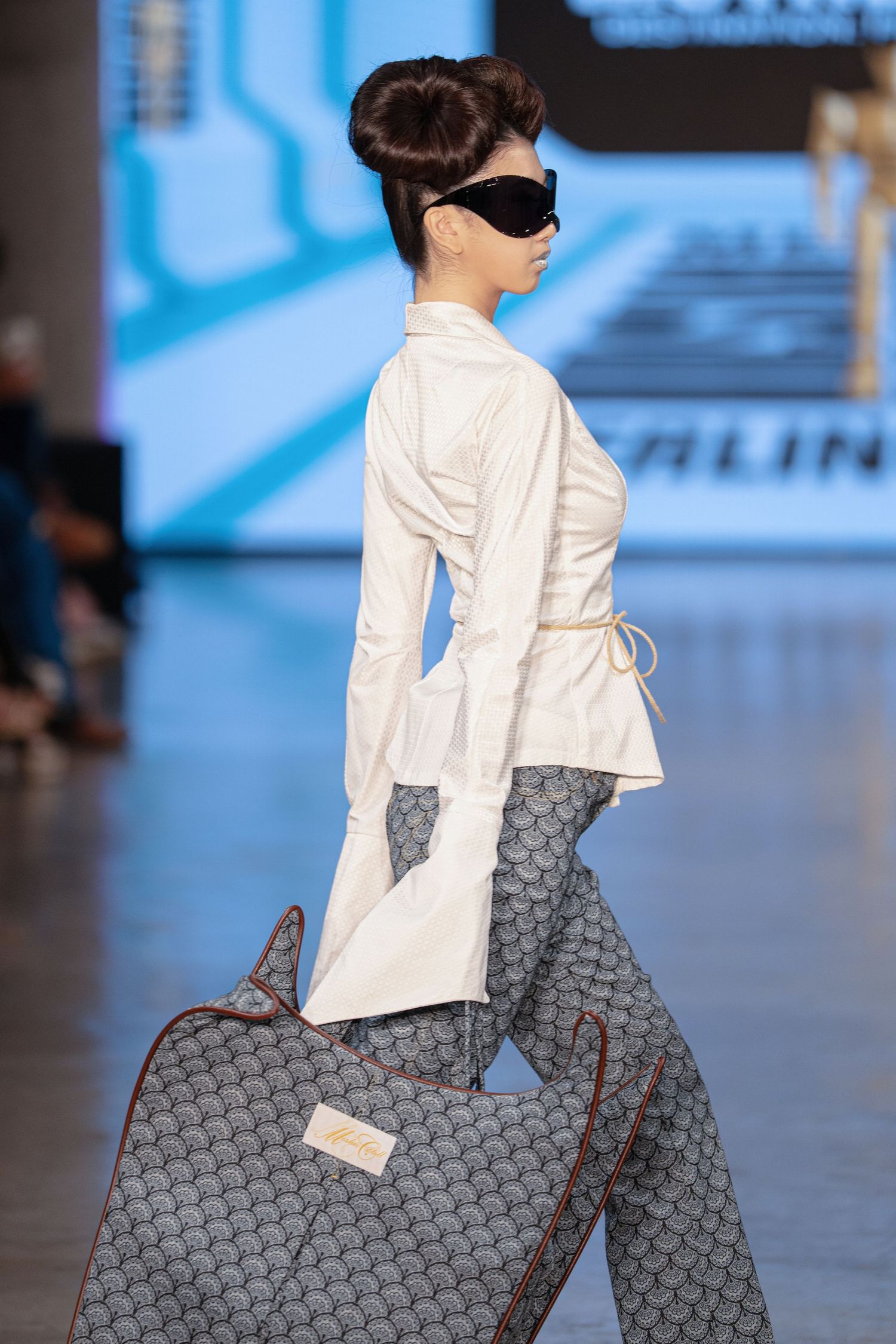 Fashion model walks runway carrying large gray patterned designer tote bag with white blouse and sunglasses.