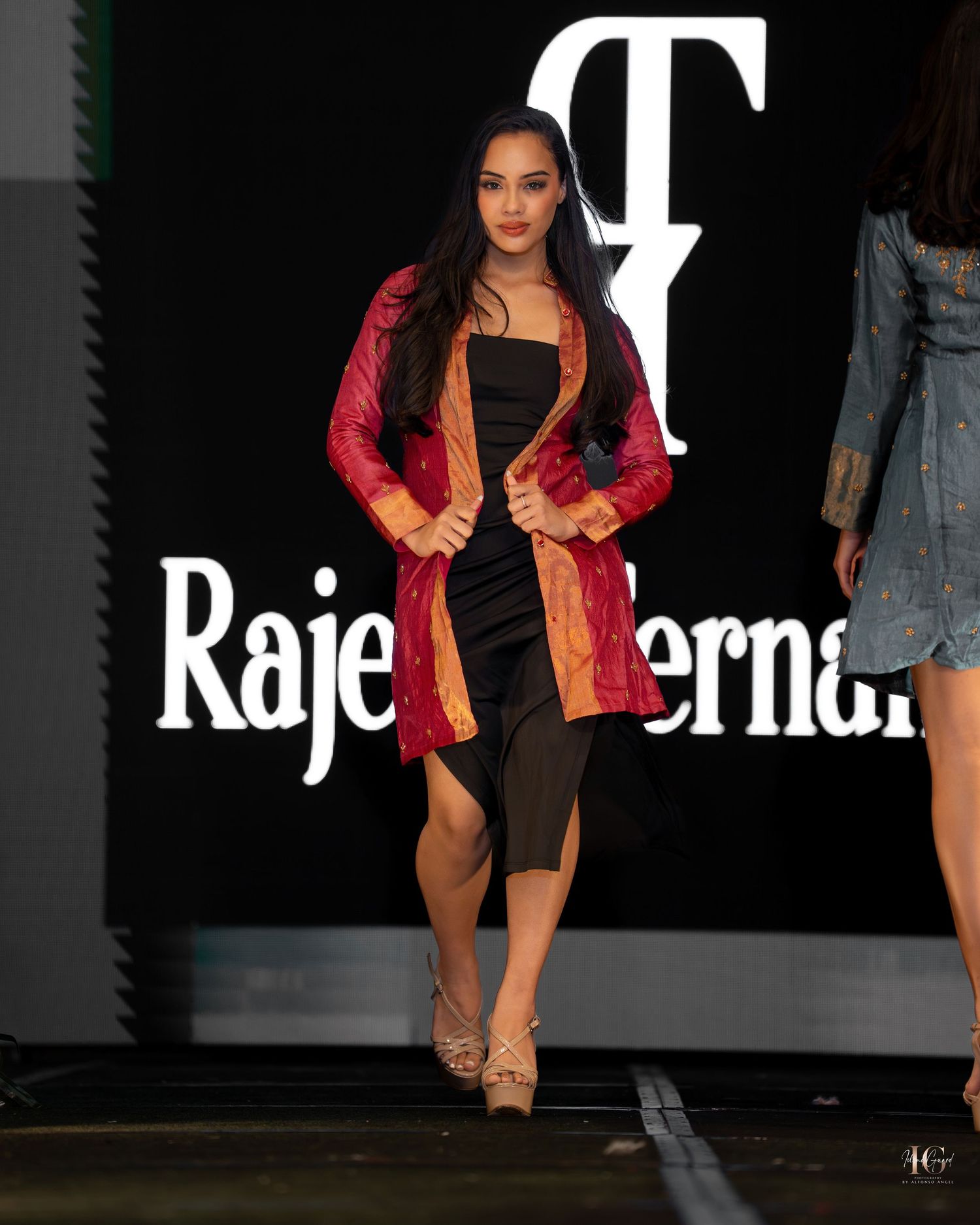 Model walks runway in red blazer and black shorts during fashion show on dark stage.