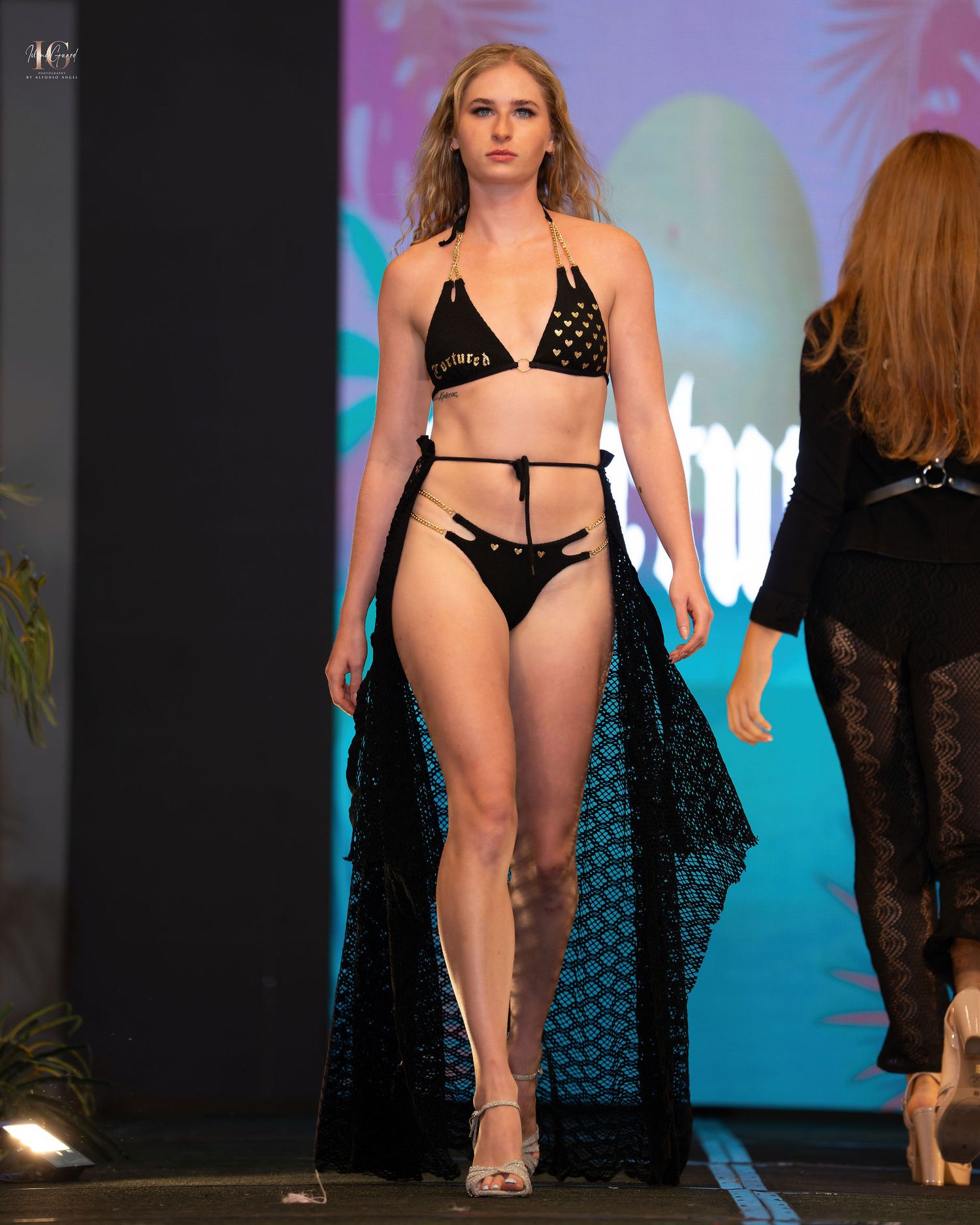 Model walks runway in black strappy bikini and flowing cape at fashion show with colorful backdrop.