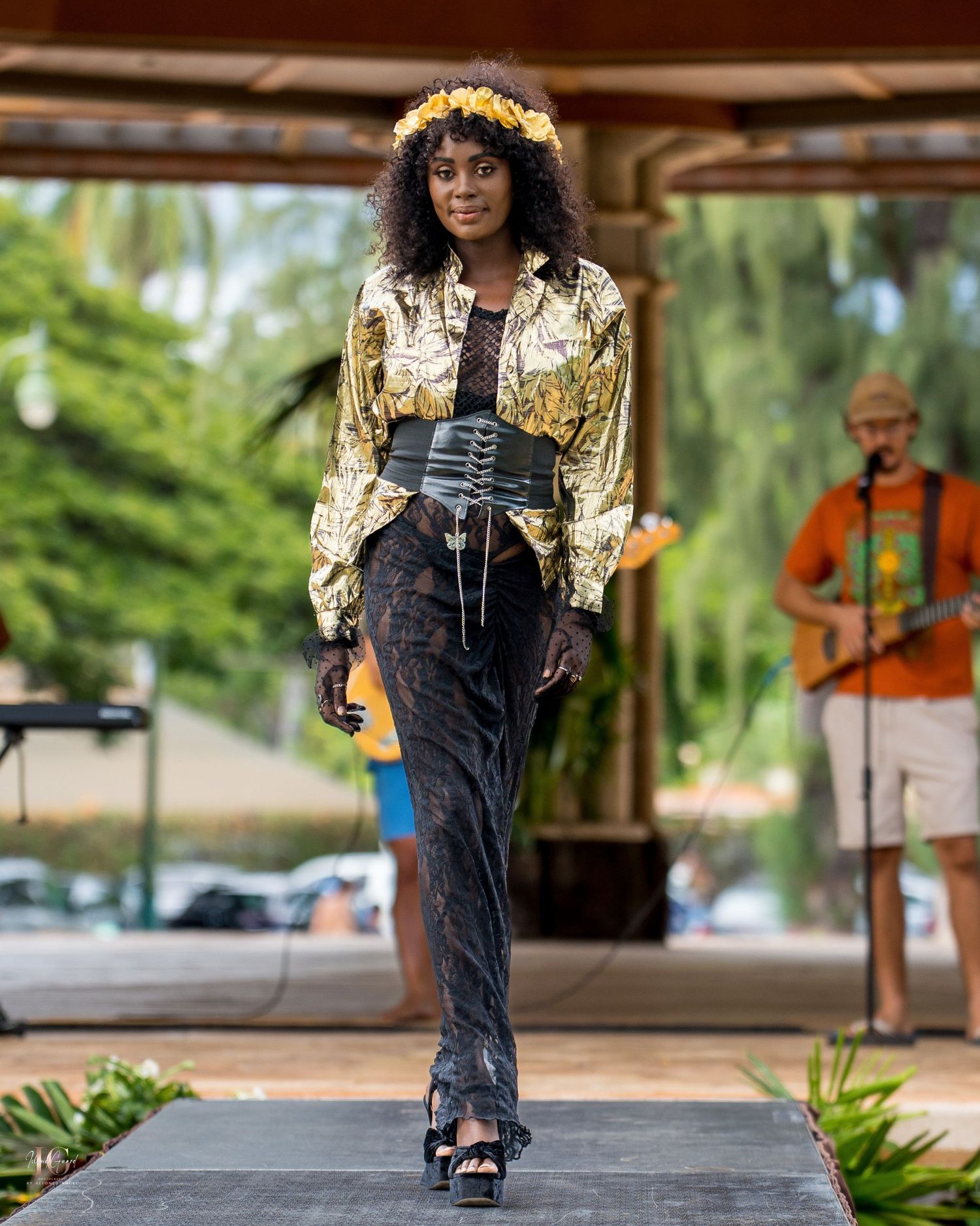 Model walks runway in denim overalls and gold metallic jacket during outdoor fashion show presentation.