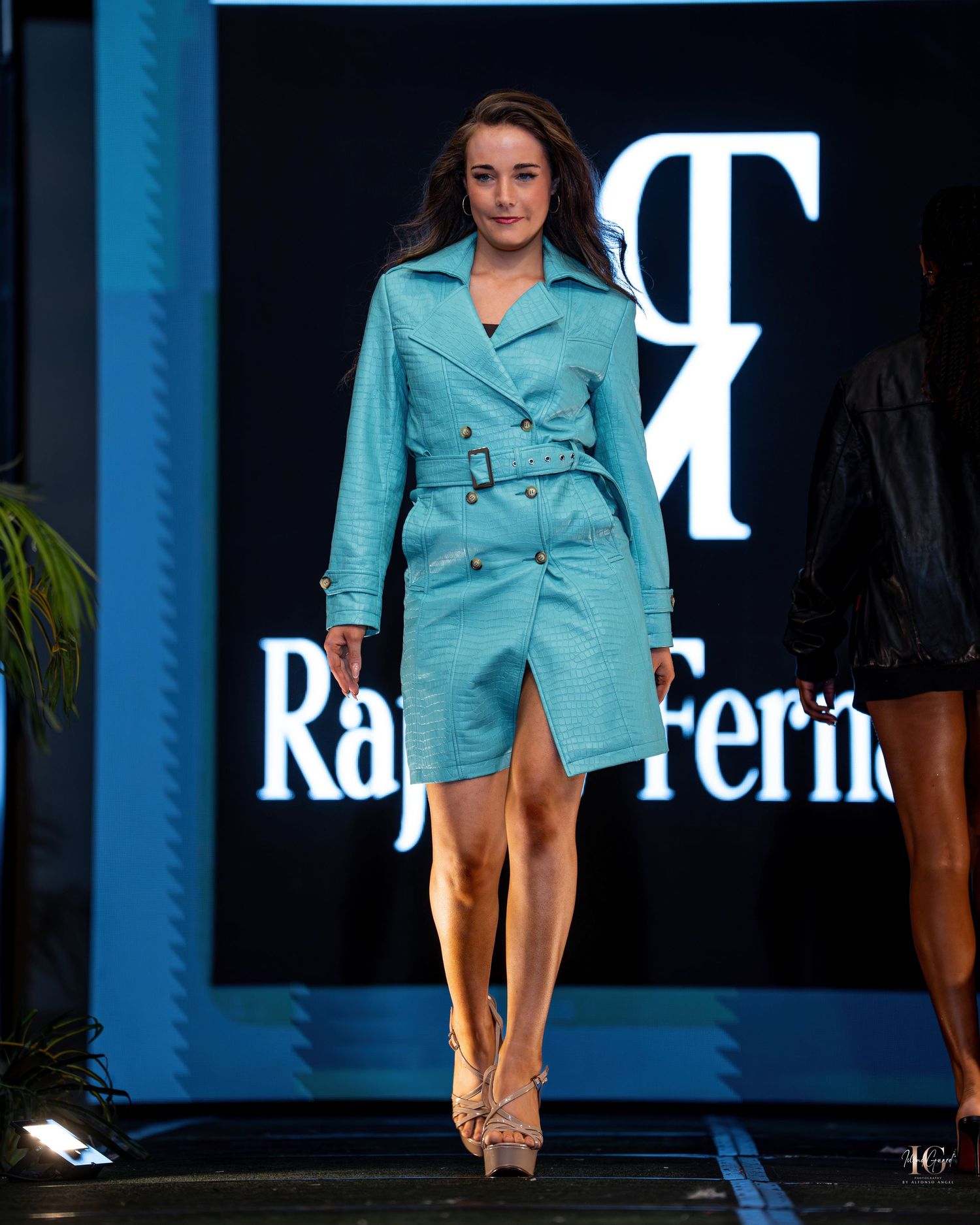 Model walks down a runway wearing a stylish turquoise trench coat dress at a fashion show.