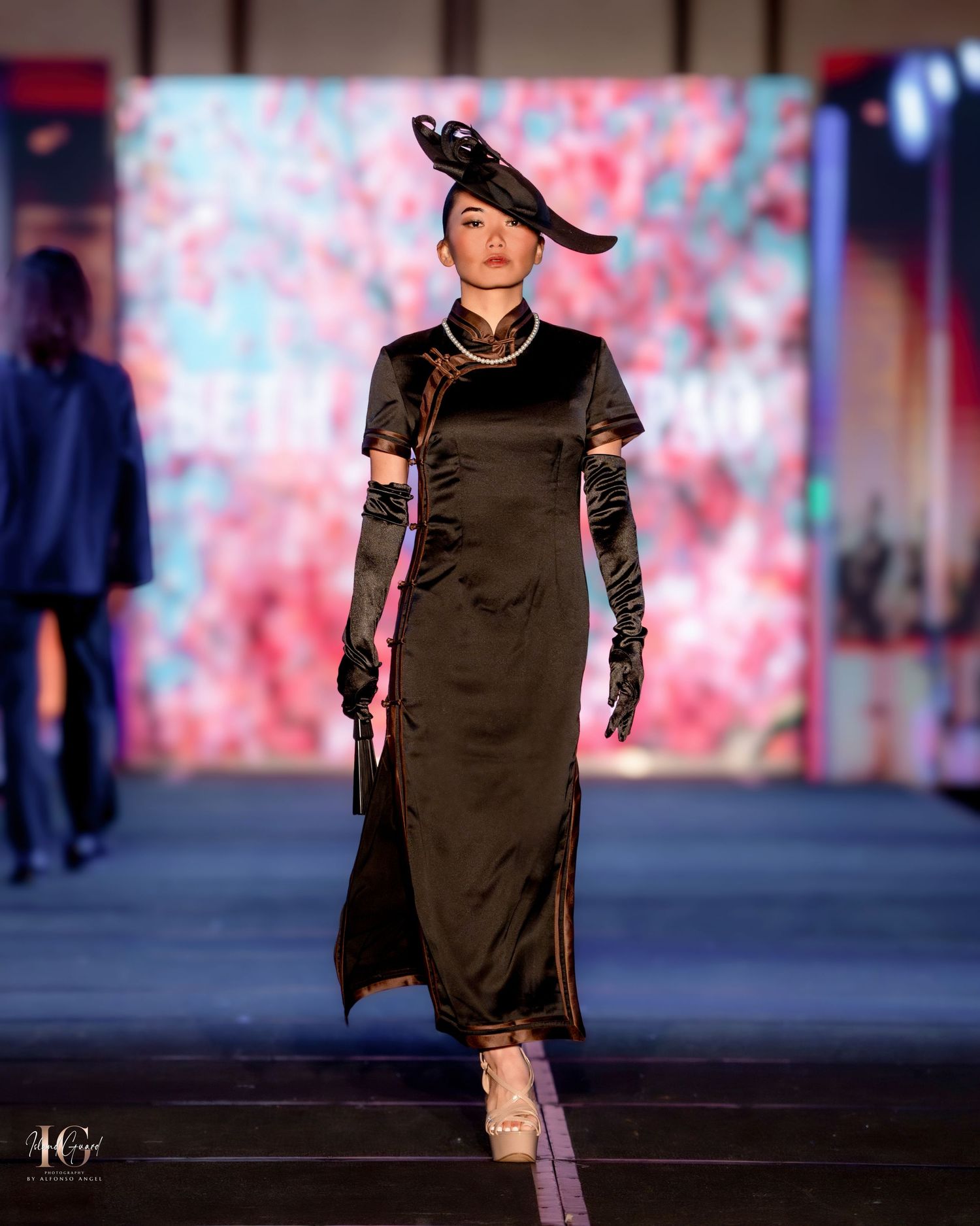 Contemporary black dress design with high neckline displayed on runway with pink backdrop.