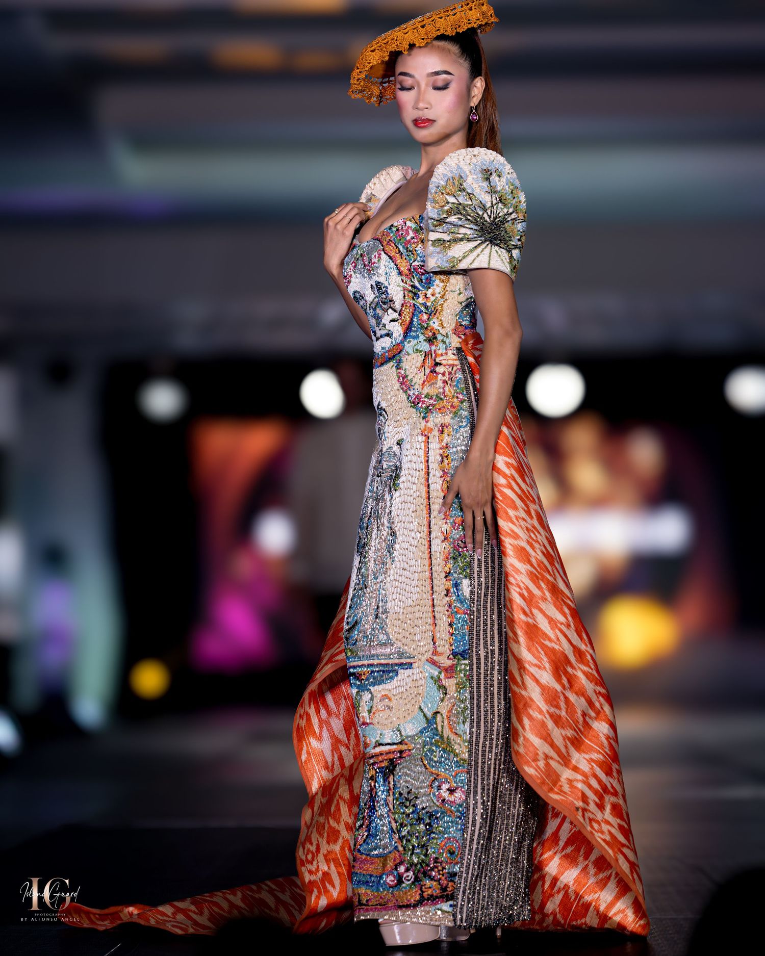 Designer gown featuring intricate patterns and orange cape flows elegantly on fashion runway.