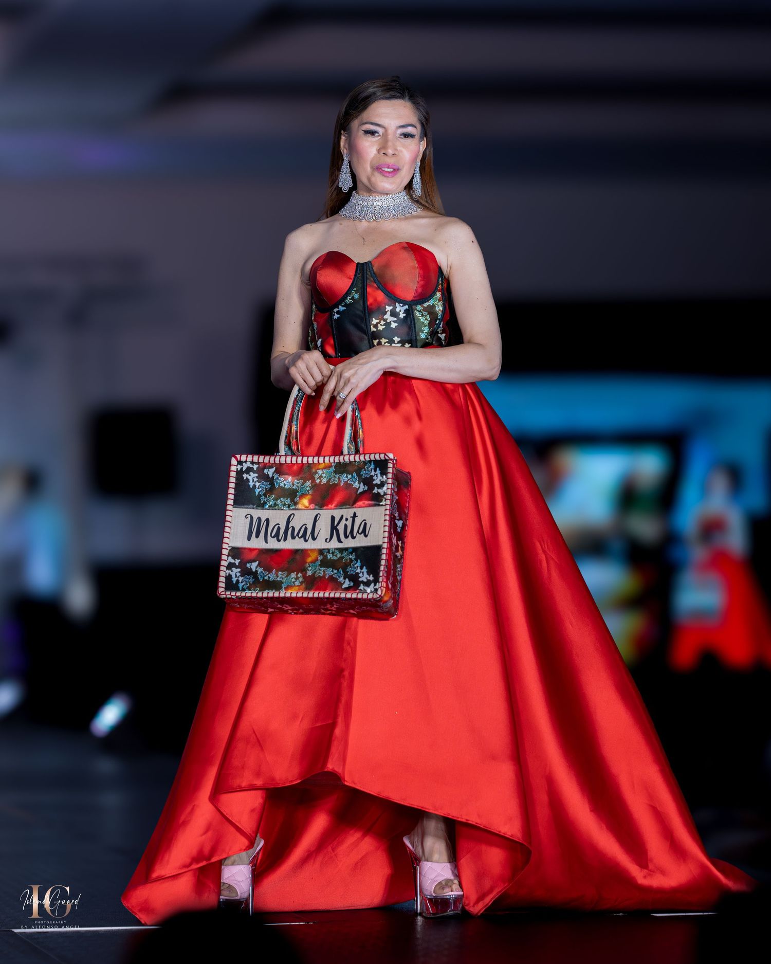 Model in dramatic red ball gown displays designer name card on illuminated catwalk.