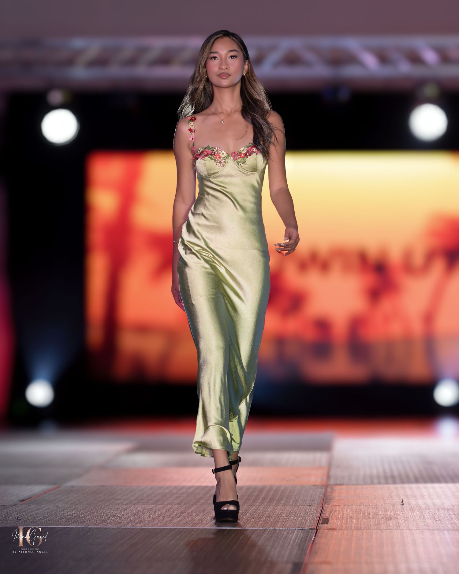 Model presents shimmering gold silk dress on fashion runway with dramatic lighting.