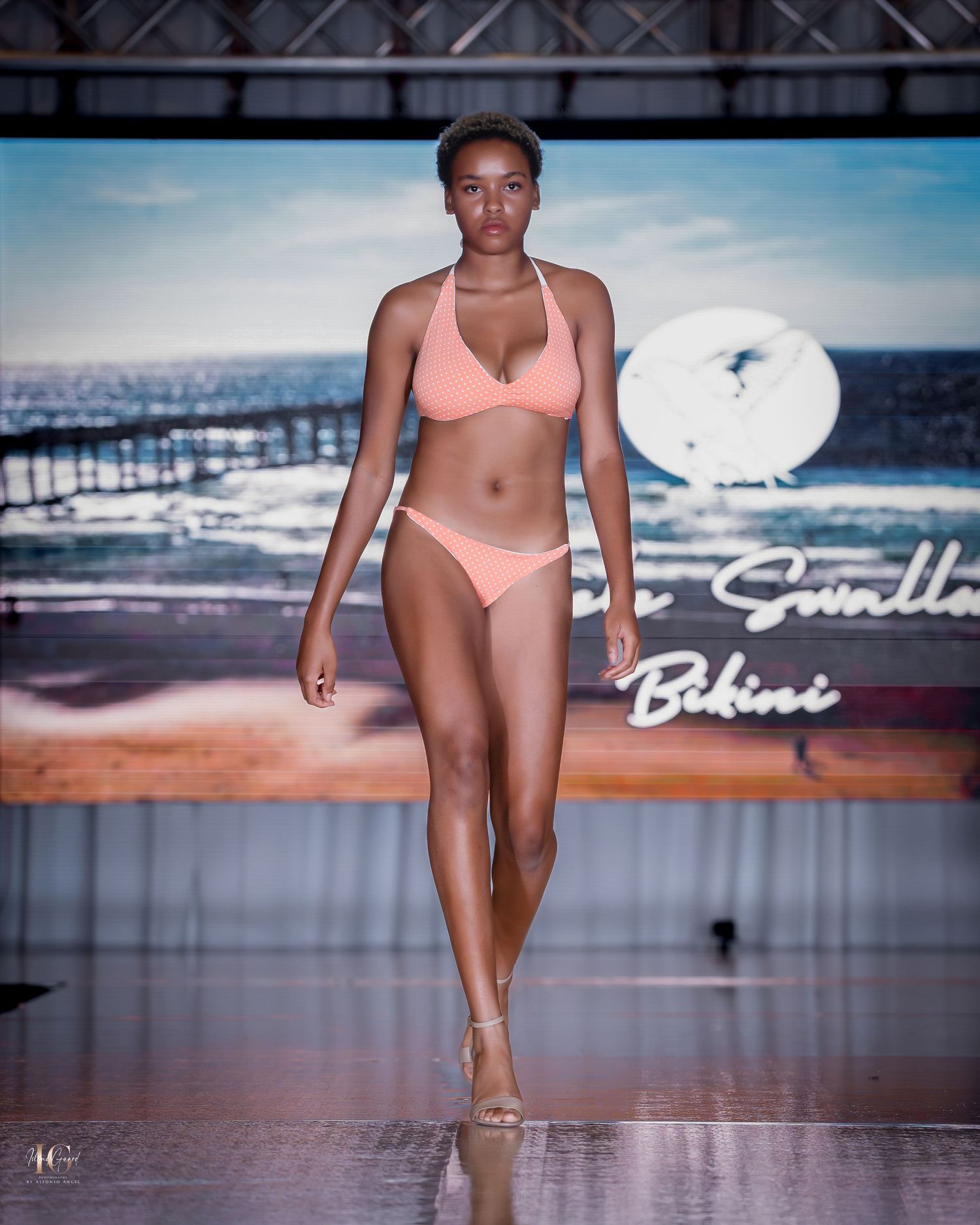 Swimwear models showcasing coral bikini designs on beachside-themed runway.