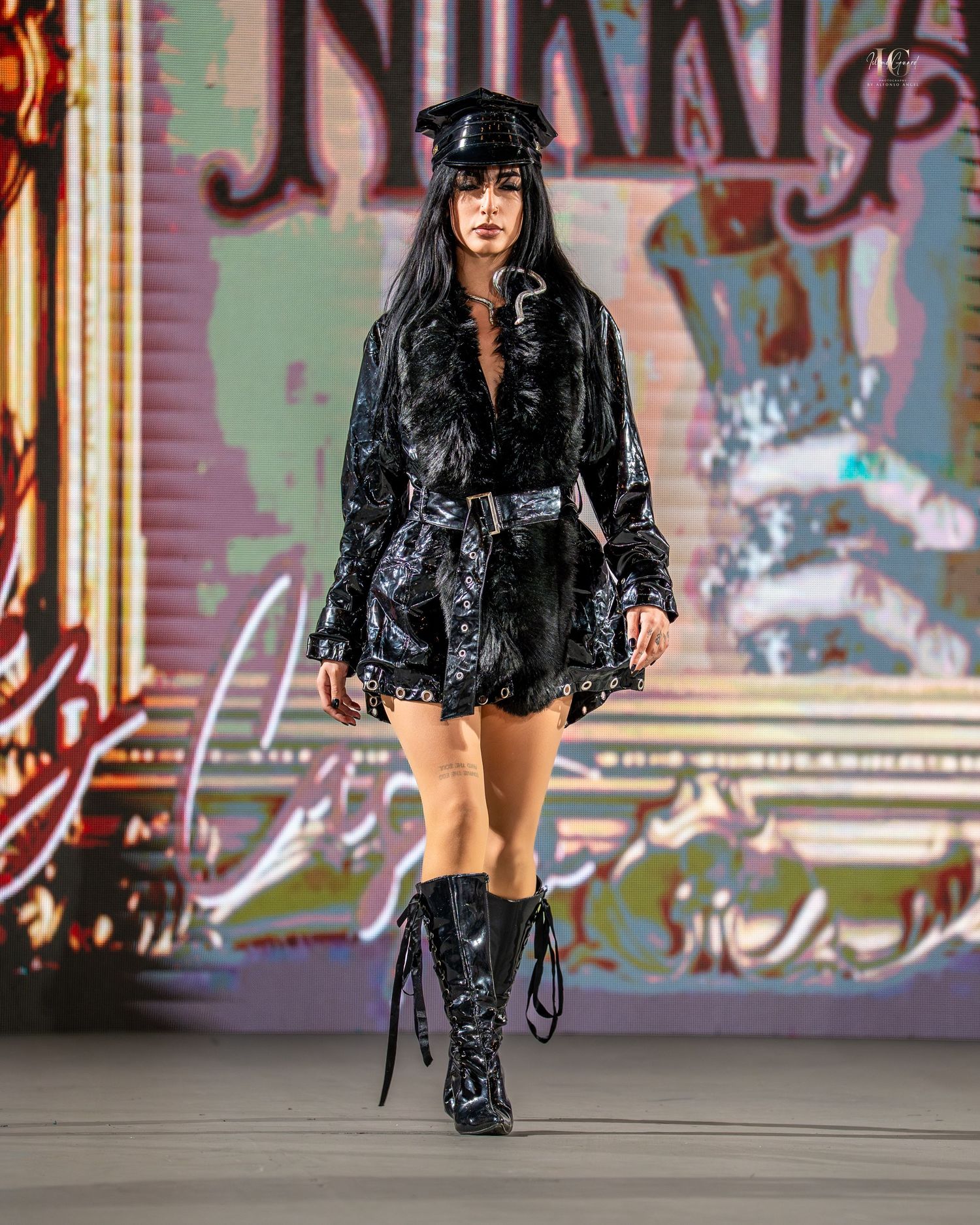 Fashion model wearing black velvet romper with matching knee-high boots and cap on runway with colorful backdrop.