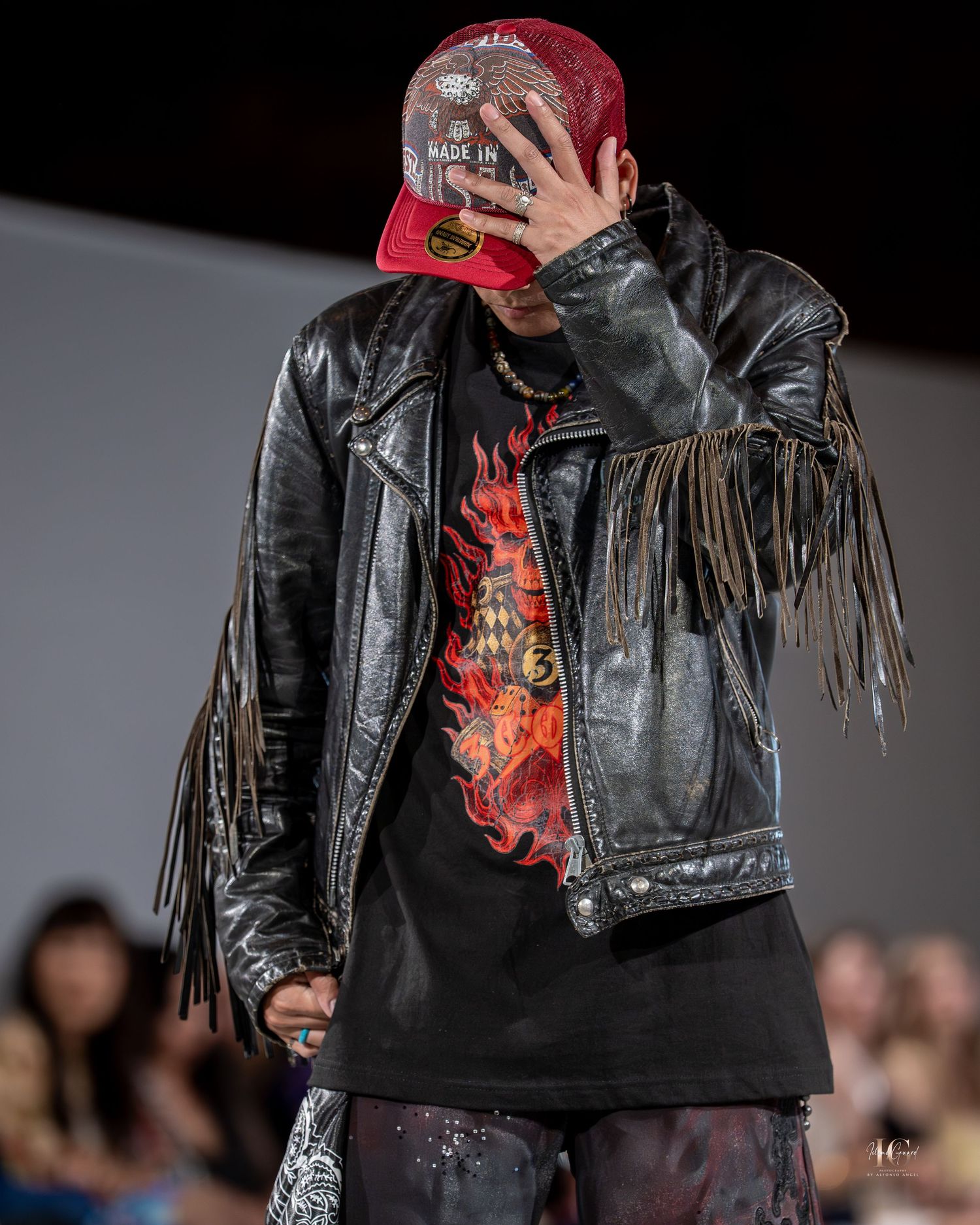 Model wearing a black leather jacket with fringe details and red accents walks the runway during fashion show.