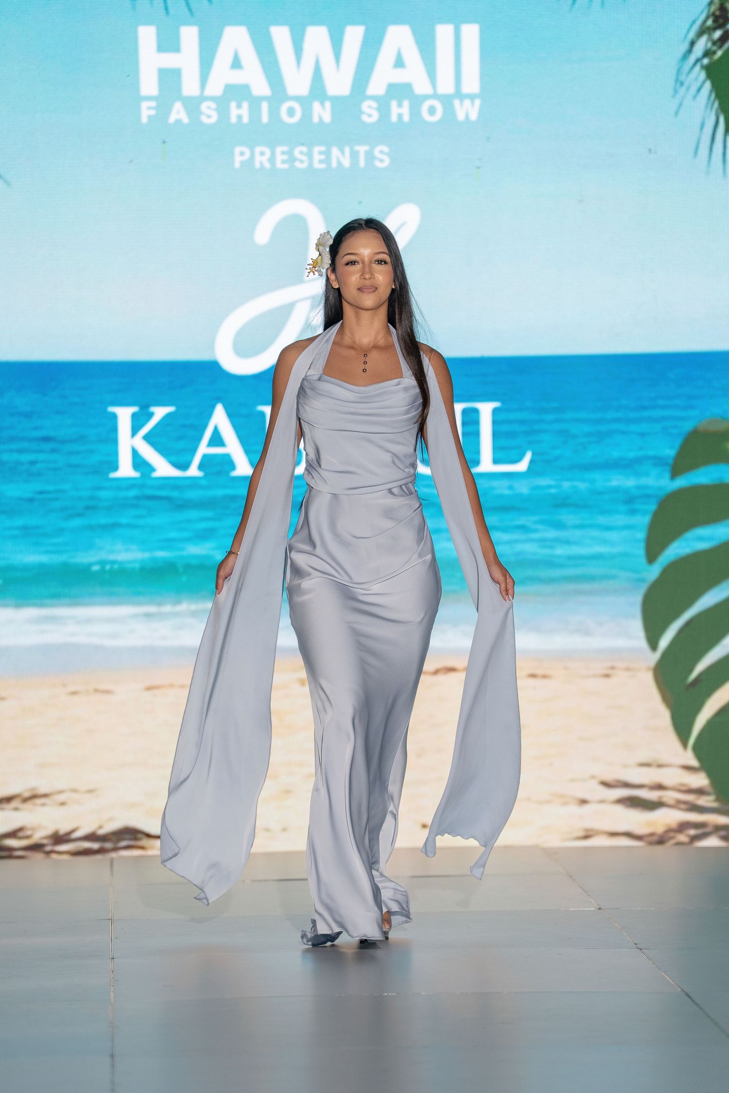 Model walks runway in flowing silver ensemble at Hawaii Fashion Show against tropical beach backdrop.