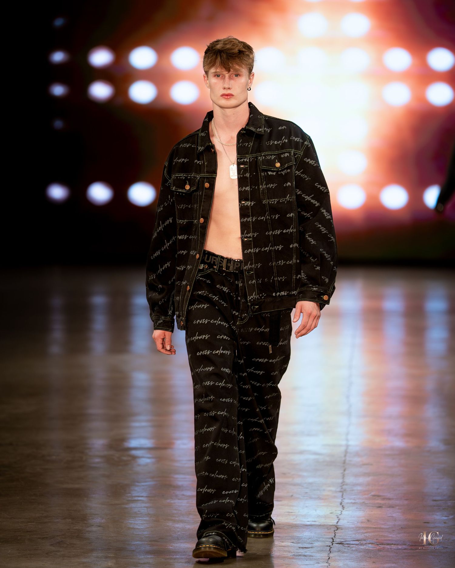 Model walks runway in black designer outfit with text print pattern under dramatic stage lighting.