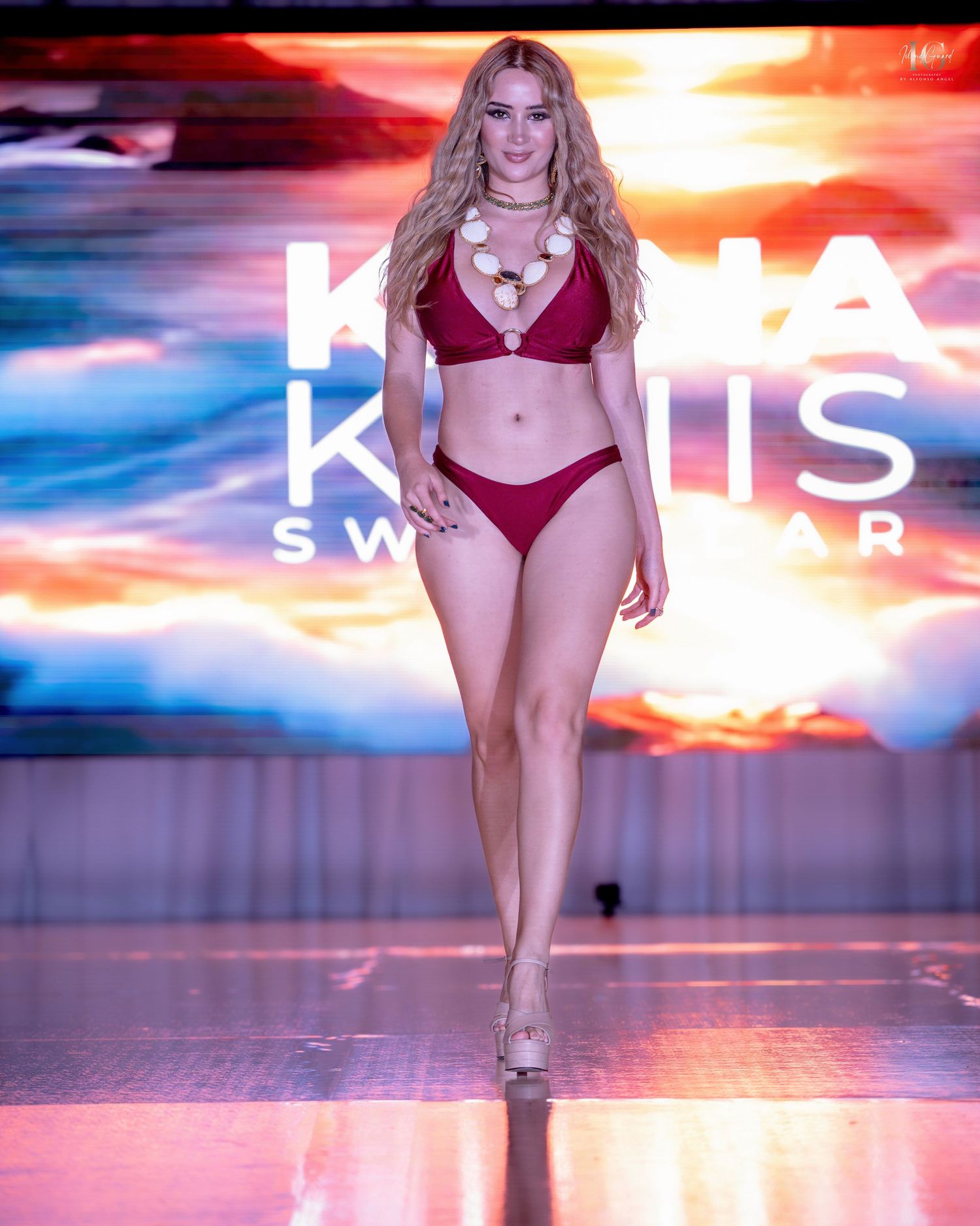 Model walks runway in red lingerie set during fashion show with dramatic sunset-colored lighting effects.