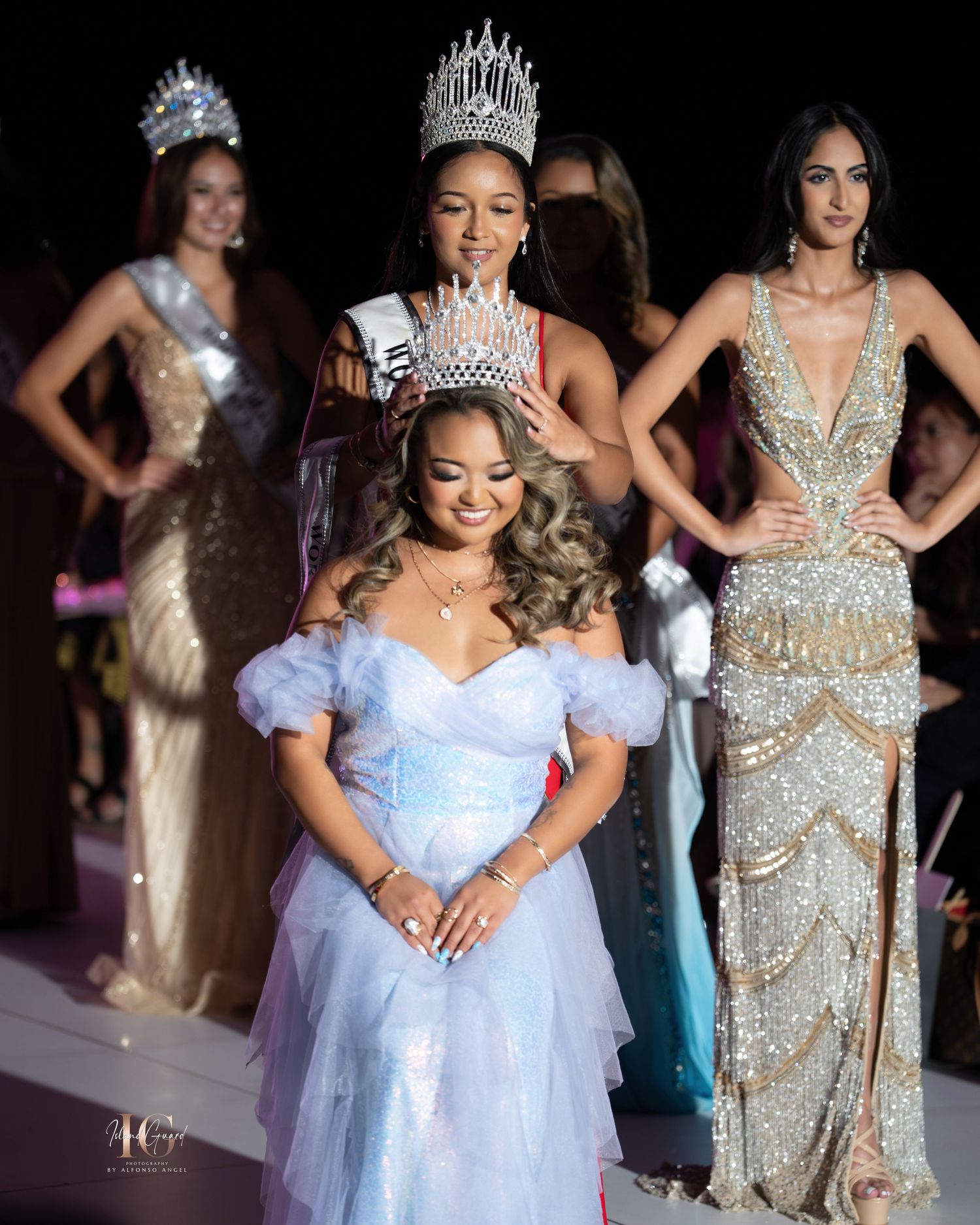 Pageant contestants in elegant evening gowns and tiaras stand together on stage during a beauty competition event.