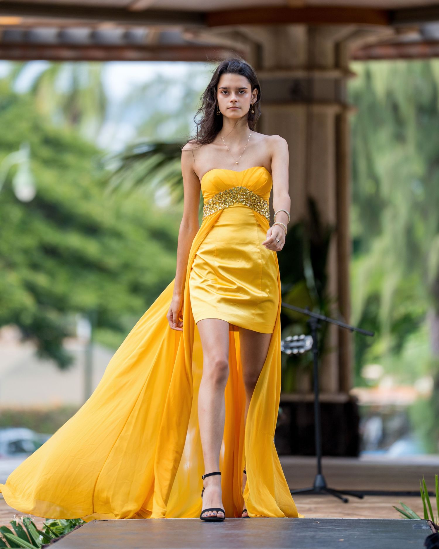 A flowing yellow evening gown with a dramatic skirt billows in the breeze against an outdoor architectural backdrop.