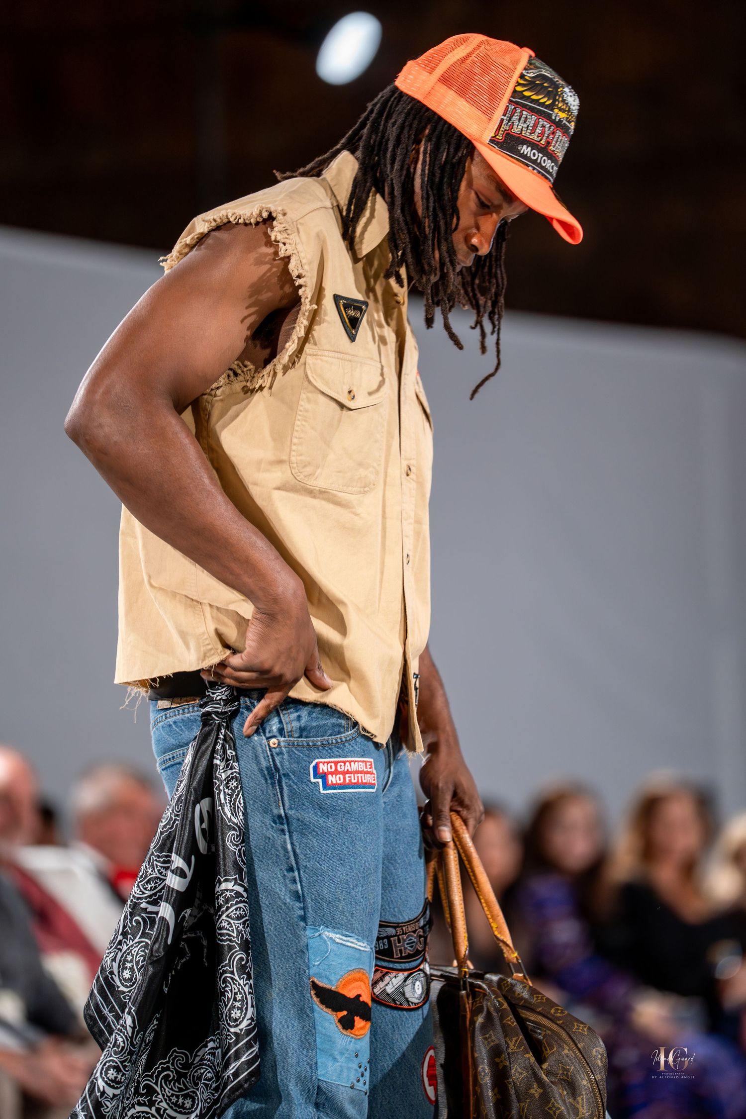Fashion model walks runway wearing tan shirt and distressed jeans with patterned bandana accessory.